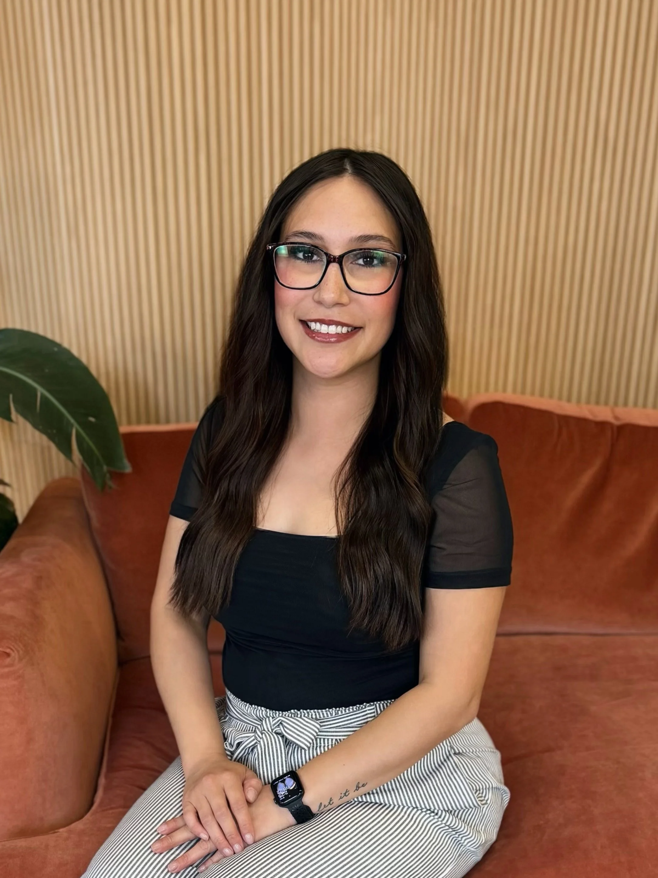 A woman with long dark hair, glasses, and tattoos, sitting on an orange velvet couch in a room with wooden panel walls and a potted plant, smiling and resting her chin on her hand.