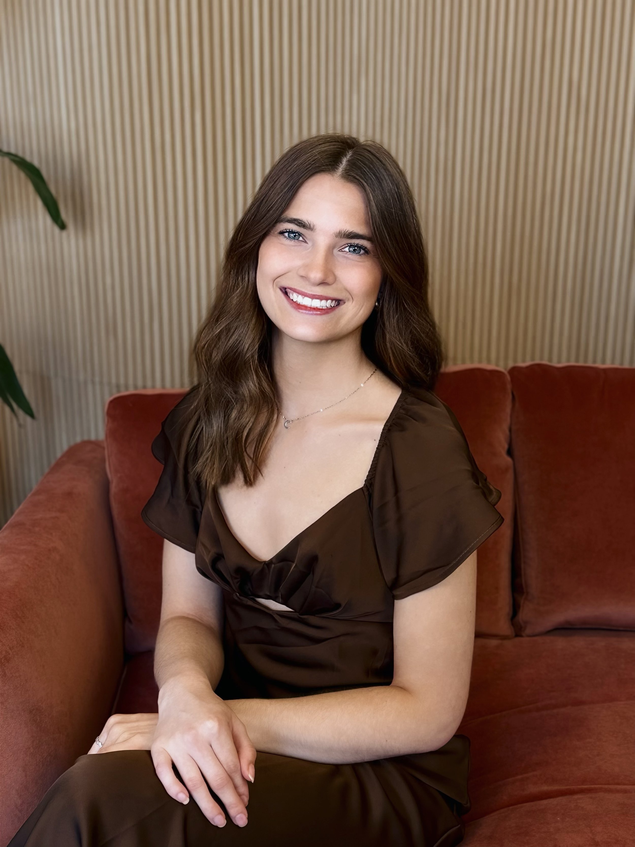 Portrait of a young woman with long wavy blonde hair, smiling, seated on a red couch in front of a wood-paneled wall, wearing a denim vest over a white long-sleeve top.