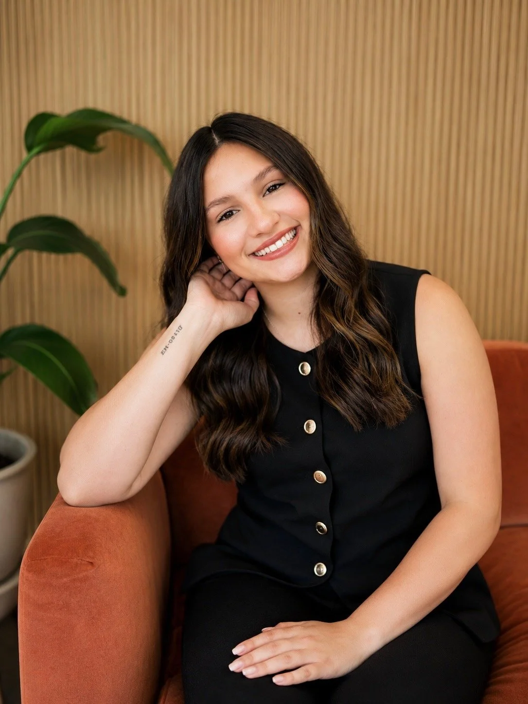 A young woman with long dark hair and a big smile, sitting on a red couch against a wood-paneled wall, wearing a light-colored long sleeve top and jeans.