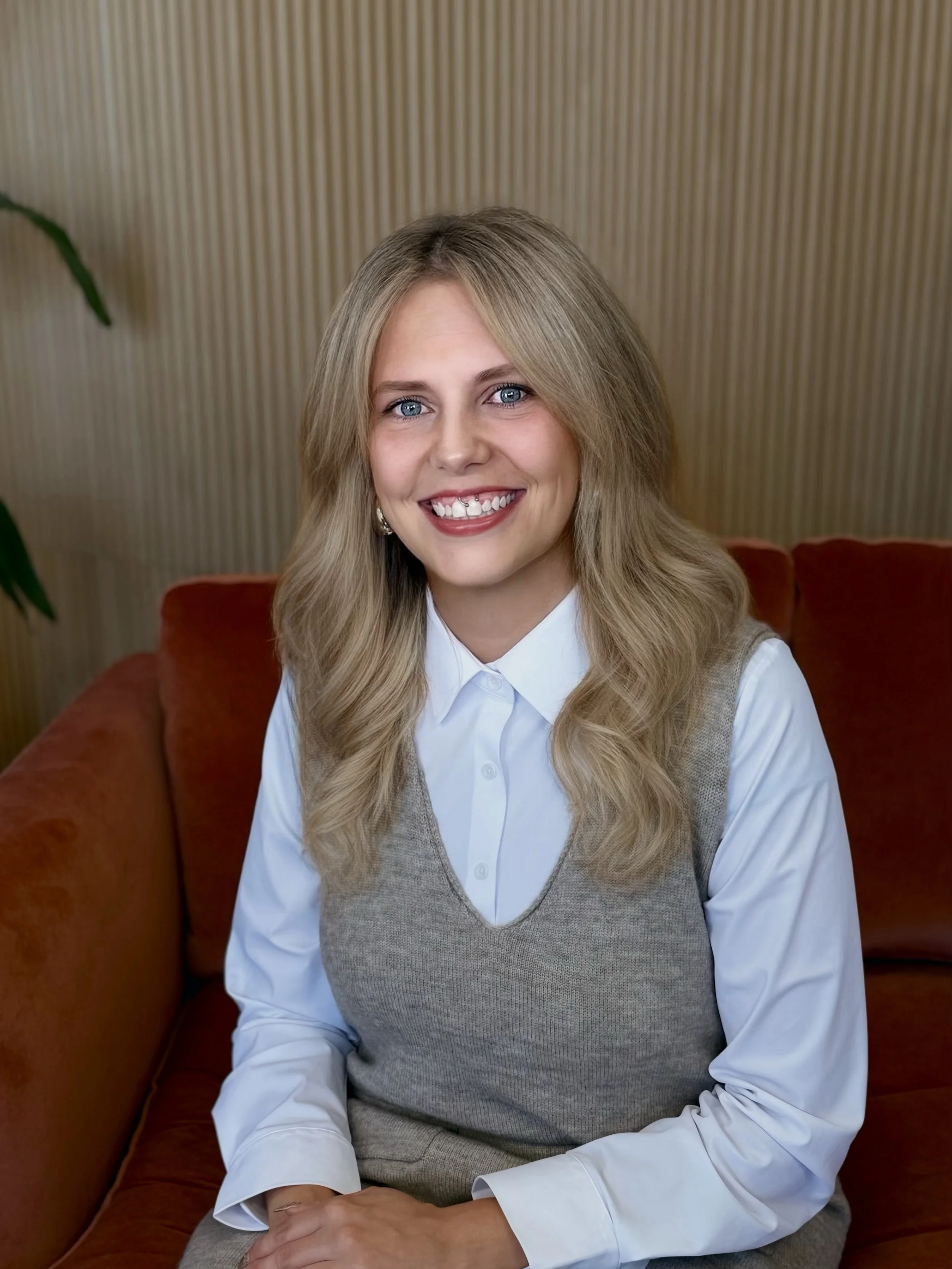 A woman with wavy blonde hair, blue eyes, and a nose piercing, smiling while resting her chin on her hand, seated indoors with a wooden-paneled background and a green plant nearby.