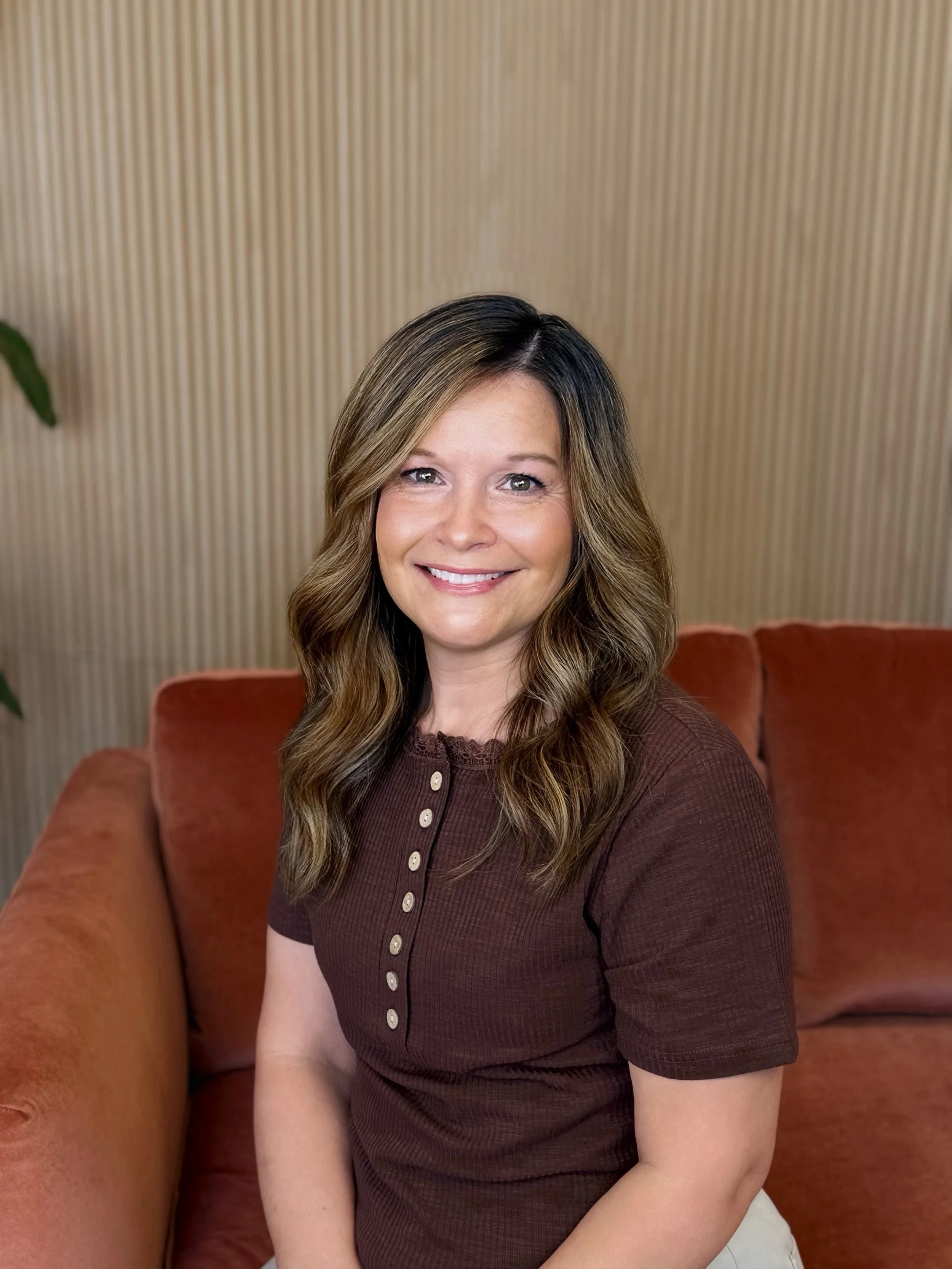A woman with shoulder-length wavy brown hair, wearing a black top with sheer polka-dot sleeves, sitting on a red sofa with a wooden striped wall in the background, smiling and touching her neck.