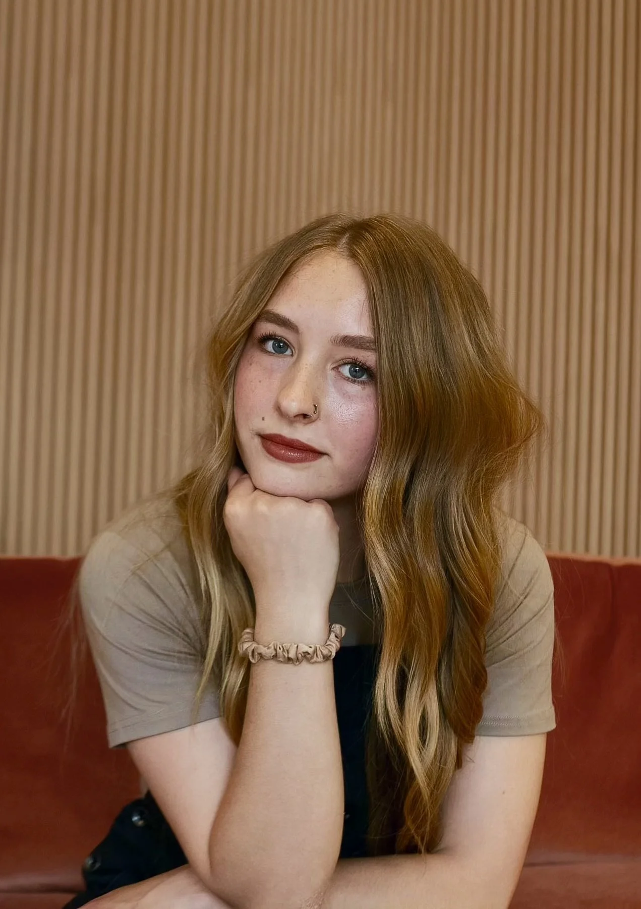 A young woman with long red hair, wearing a gray shirt and denim jacket, smiling while sitting against a wooden wall.
