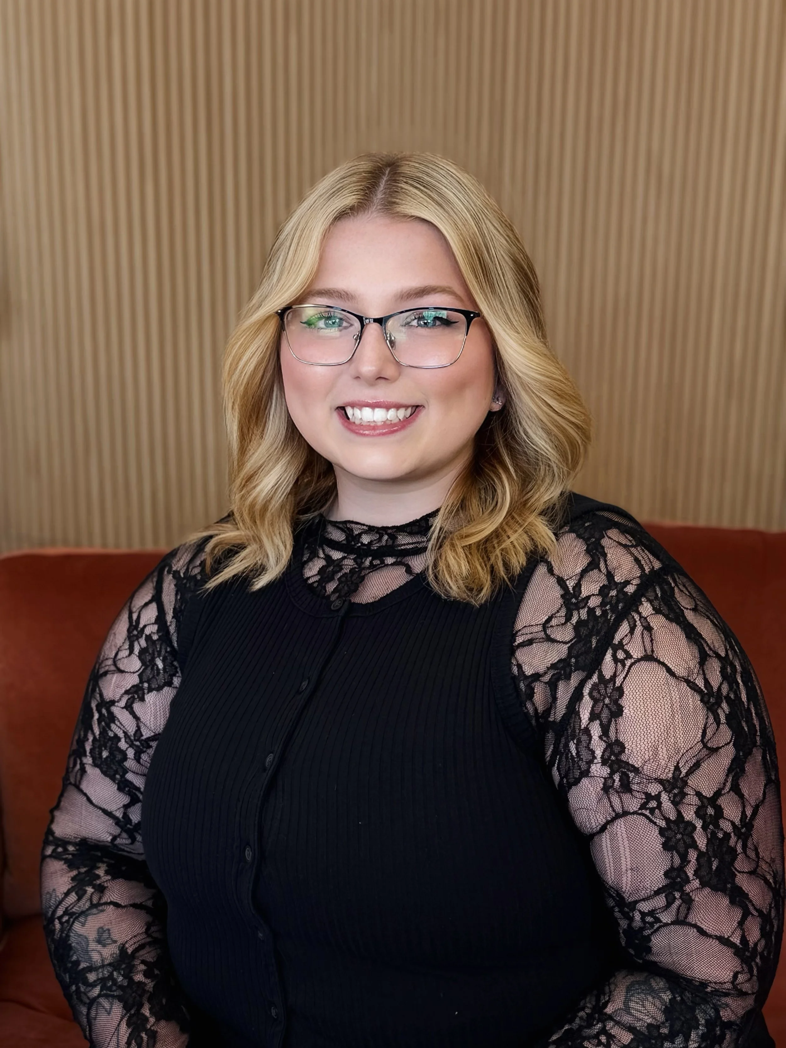 A young woman with shoulder-length brown hair, glasses, and tattoos on her arms, wearing a sleeveless black top, sitting on a wooden bench with a striped wooden wall behind her, smiling gently.