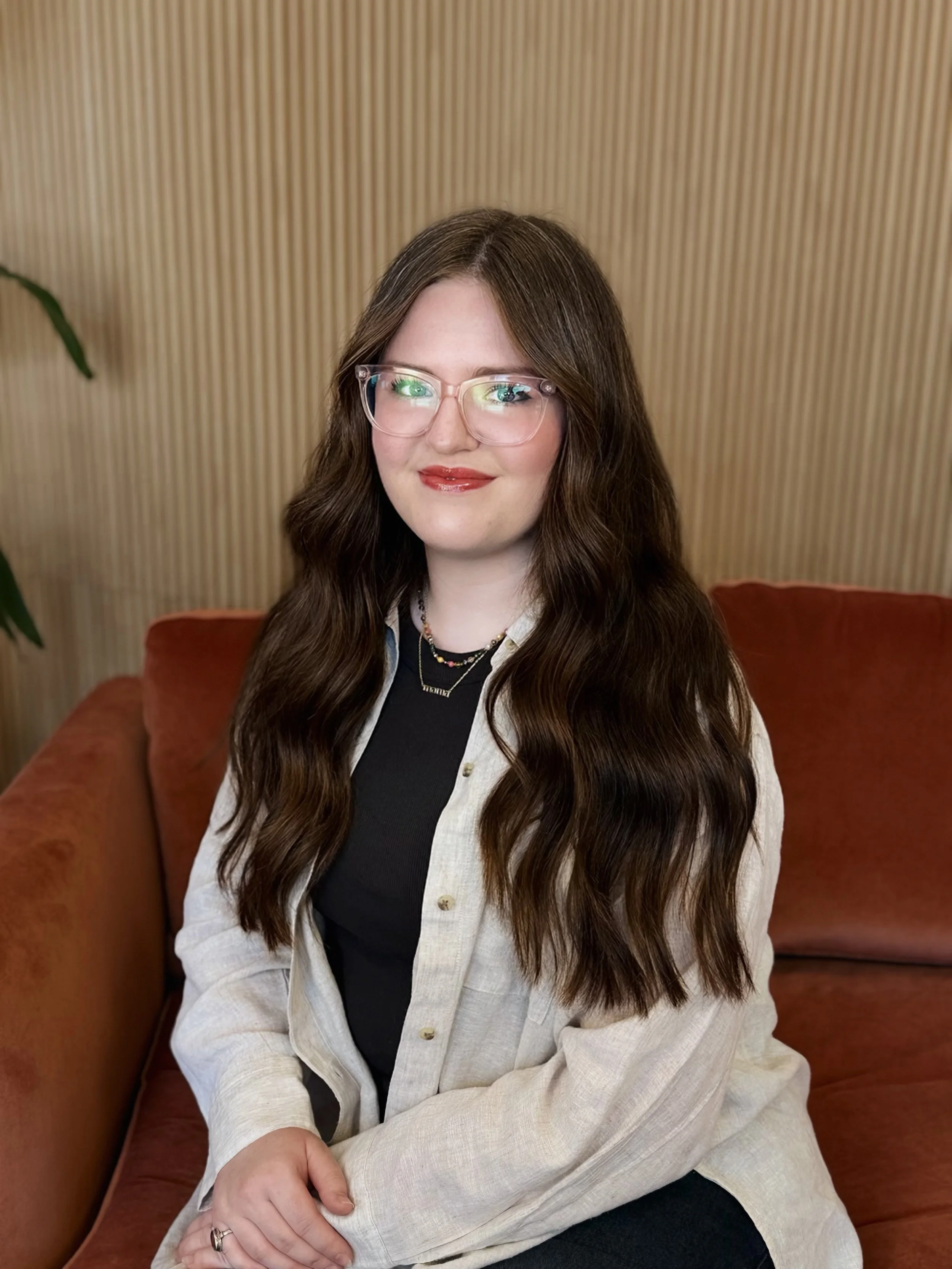 A woman with long dark wavy hair, wearing glasses, a brown top, a beige fuzzy cardigan, and blue jeans, sitting on a red velvet couch against a wooden paneled wall.