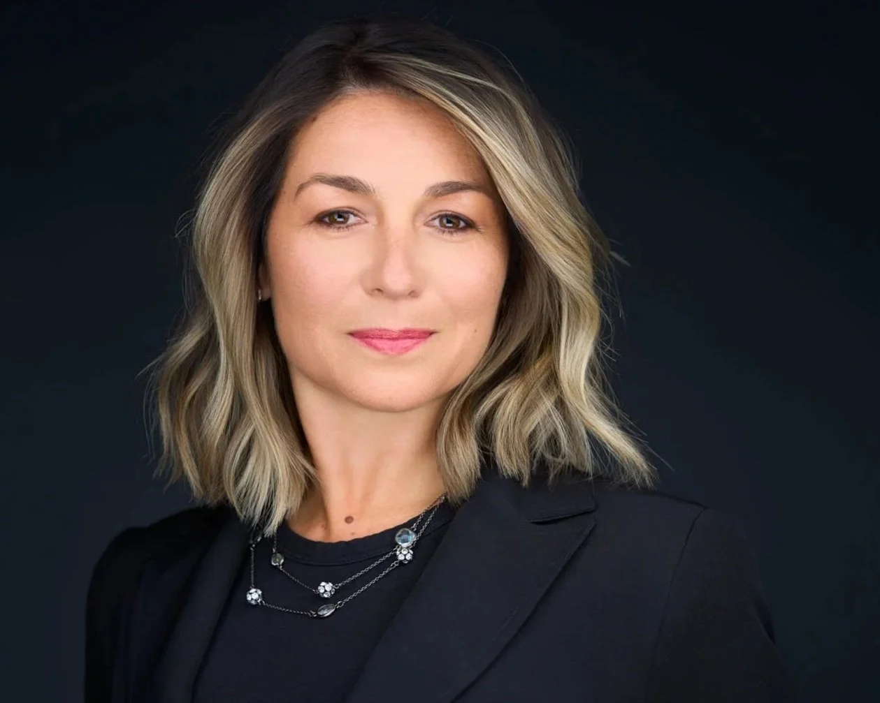 Portrait of a woman with blonde, wavy hair wearing a black blazer and layered necklaces against a dark background.