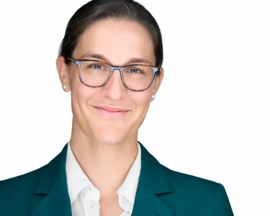 Professional woman with glasses smiling in a teal blazer and white shirt against a white background.