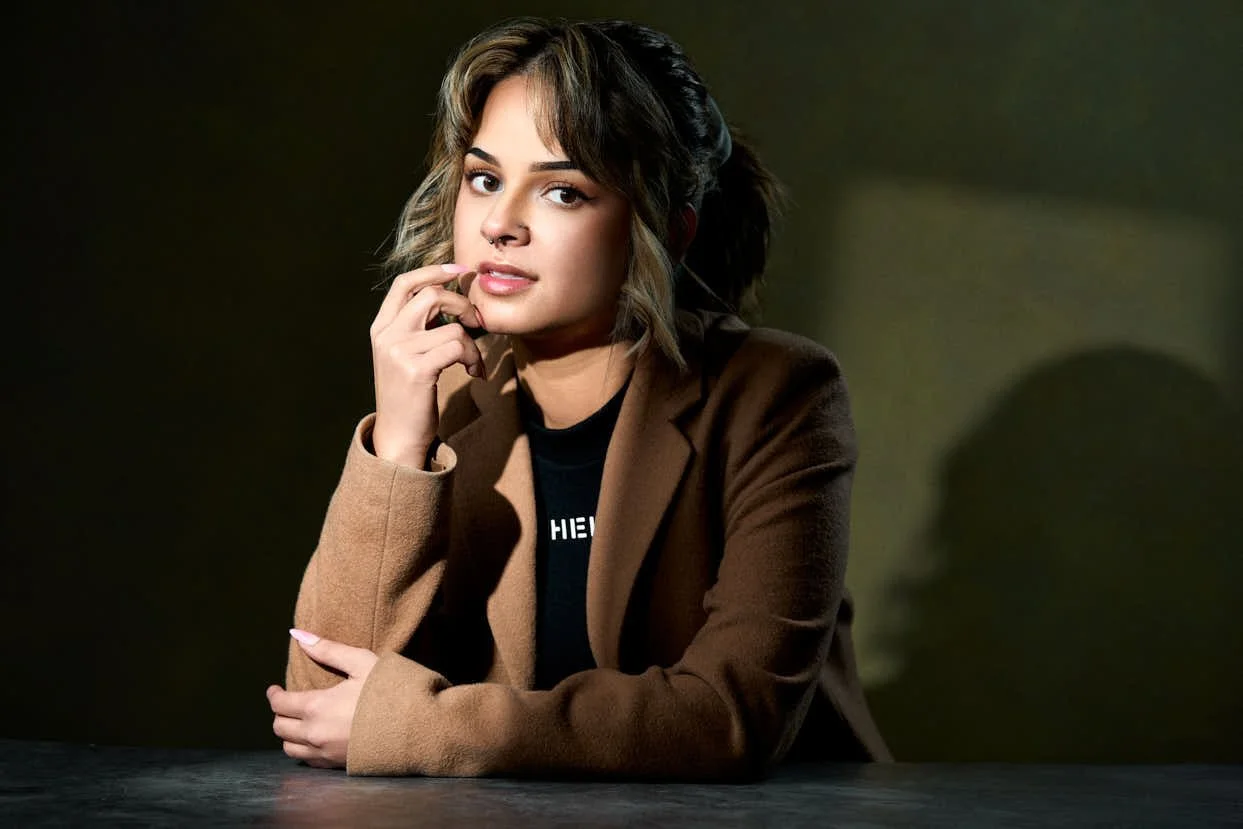 A young woman with wavy hair, wearing a brown blazer and black t-shirt, sitting at a table with a dark background.