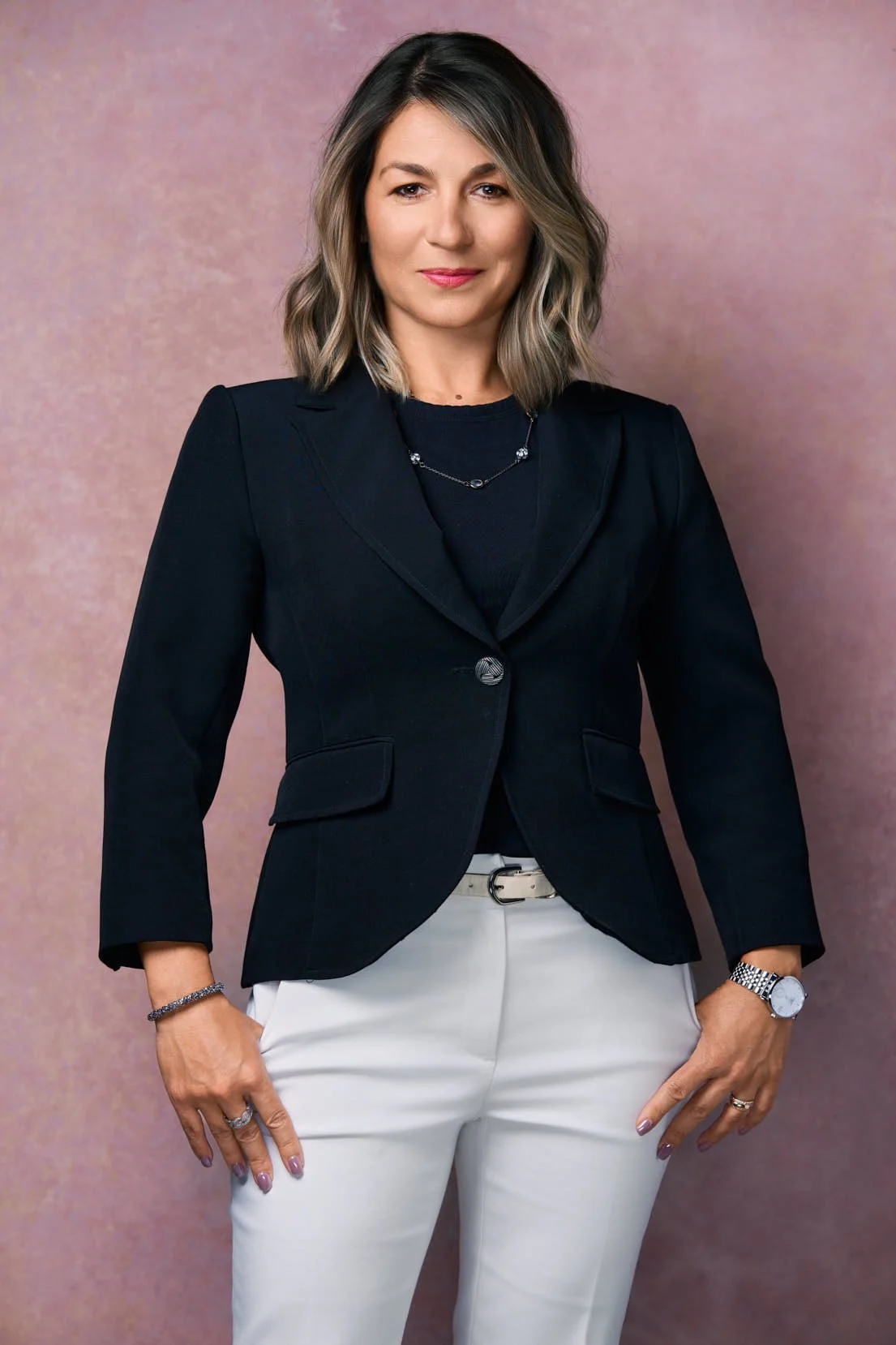 A woman with shoulder-length wavy hair wearing a black blazer, a necklace, white pants, and accessories, standing against a pink background.