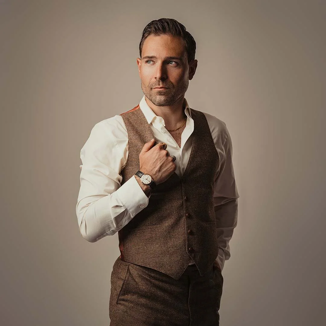 A man with dark hair and a beard wearing a white dress shirt, brown vest, and dark pants, posing against a plain background while holding his bag strap over his shoulder.