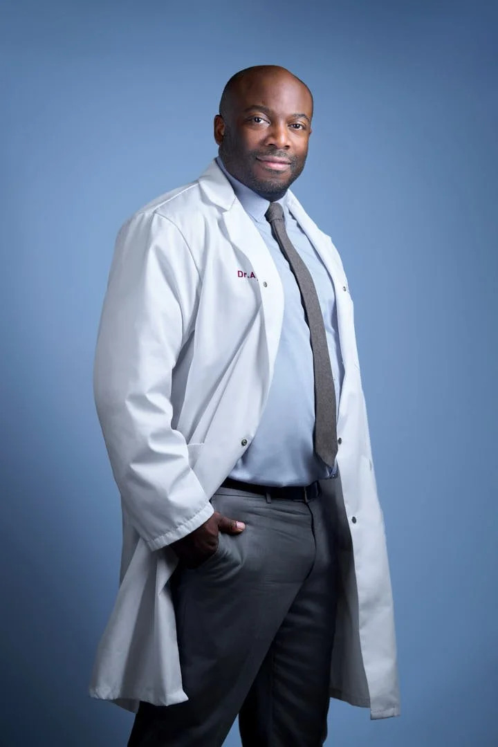 A confident African American male doctor wearing a white coat, dress shirt, and tie, standing against a blue background.