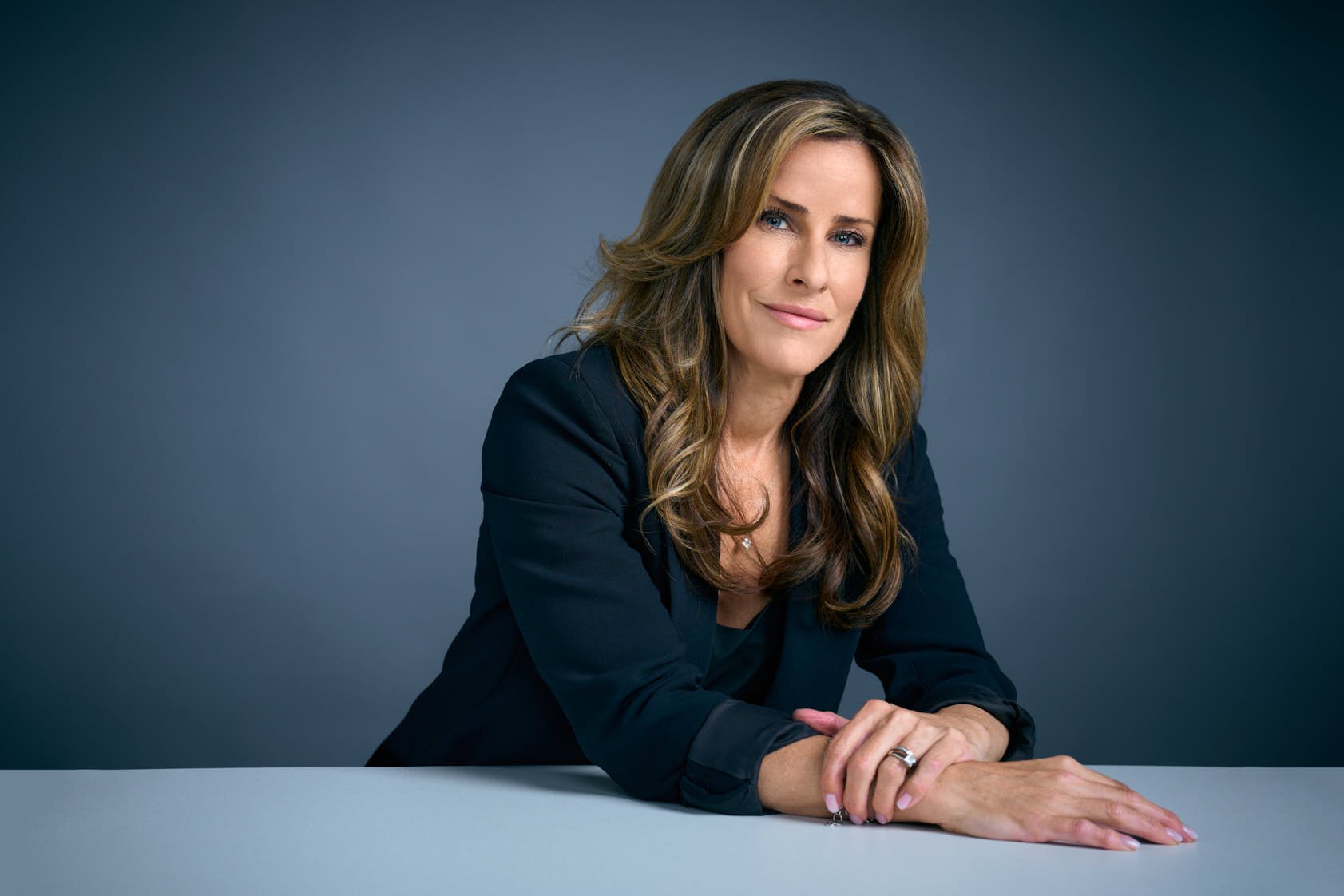 A woman with wavy brown hair, blue eyes, and a confident expression, dressed in a dark blazer, sitting at a table against a gray background.