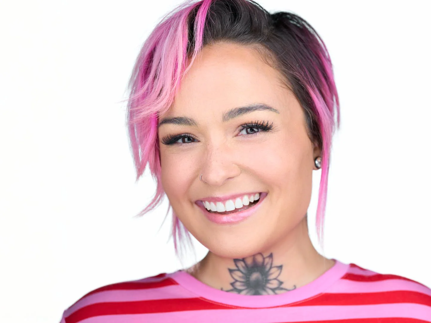 A woman with short pink and black hair, wearing a pink and red striped shirt, smiling with teeth showing, with a tattoo of a flower on her neck, earrings, and a nose ring, against a white background.