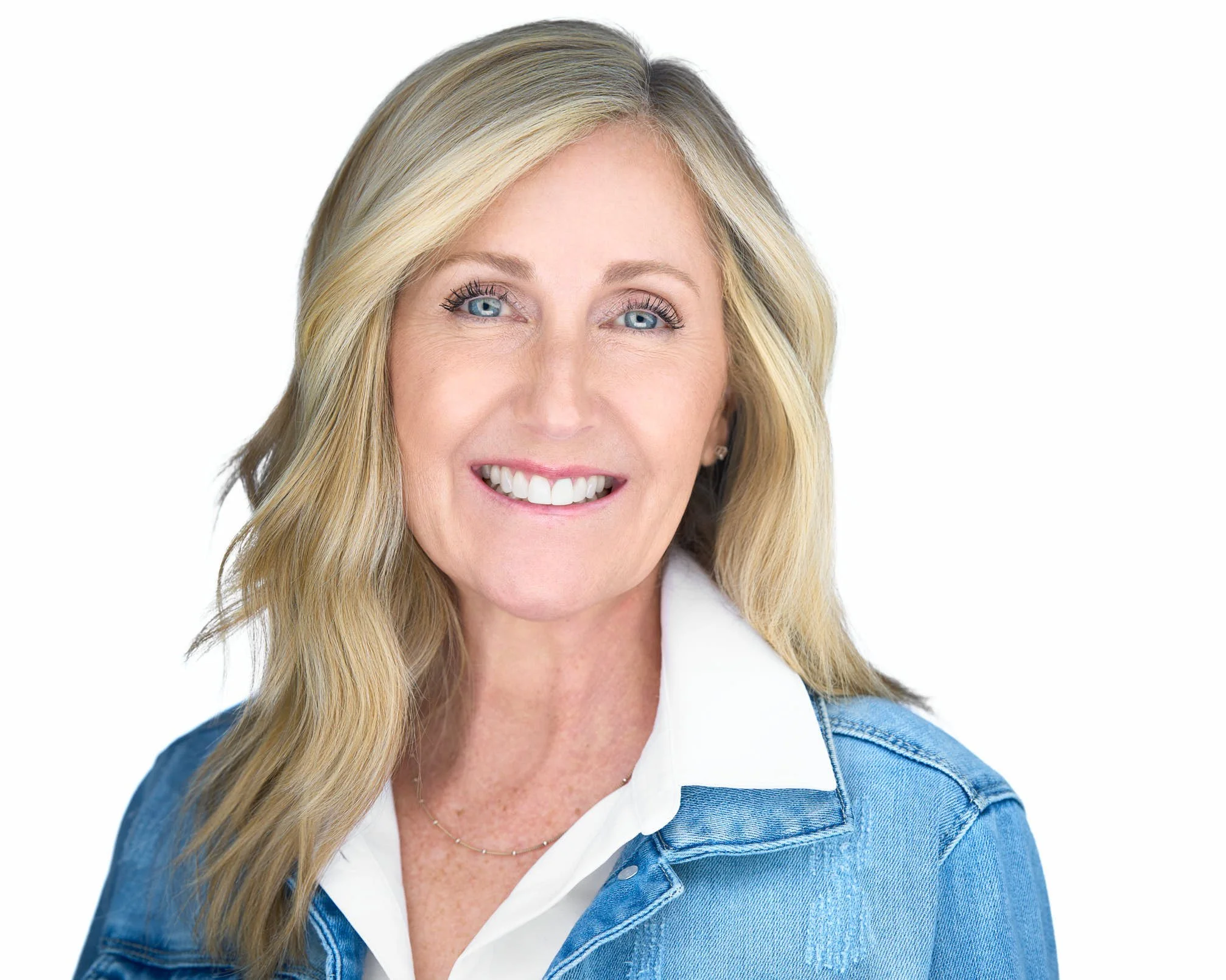 A smiling blonde woman with blue eyes wearing a white shirt and denim jacket, posing against a white background.