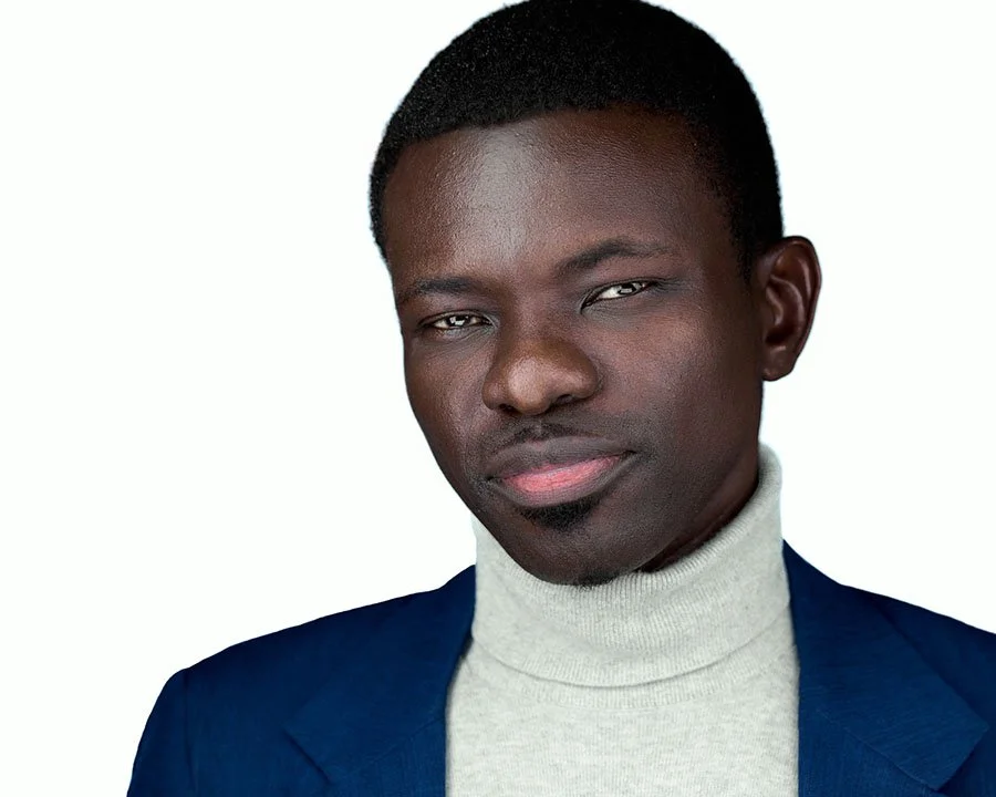 A young African American man with short hair, wearing a light gray turtleneck and a dark blue blazer, looking directly at the camera with a serious expression.