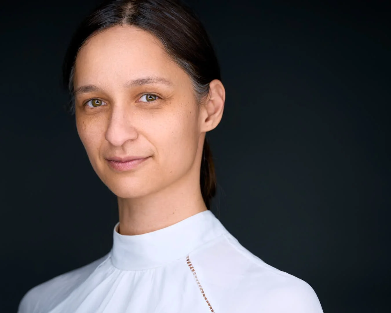 Close-up portrait of a woman with dark brown hair, fair skin, and hazel eyes, wearing a white top, against a black background.