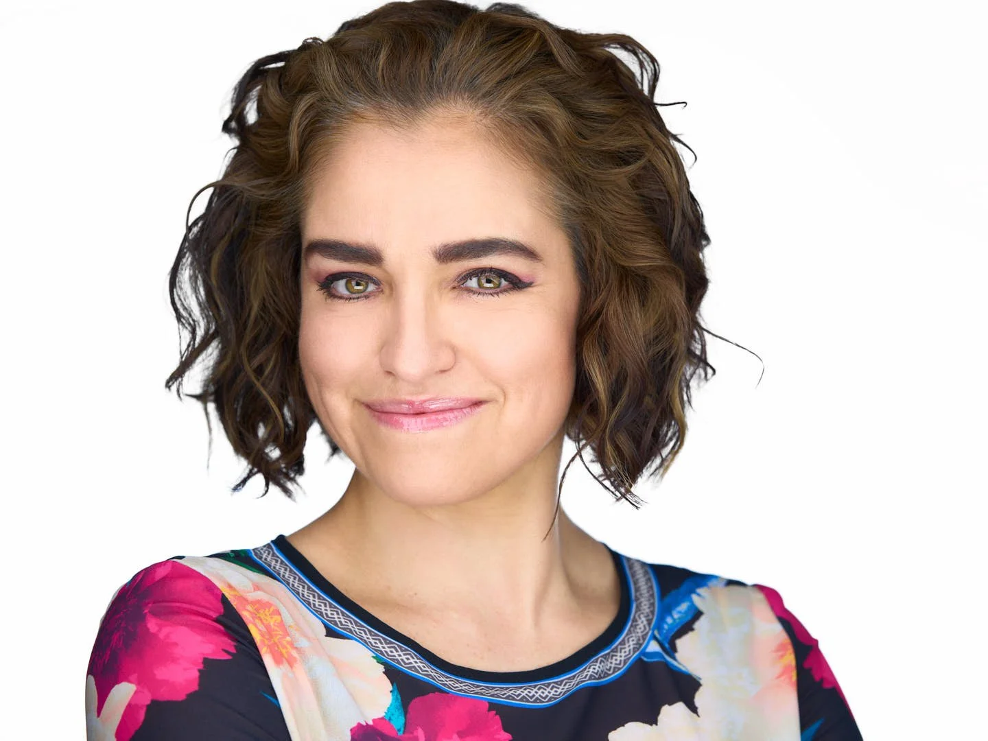 A woman with short curly brown hair, wearing a colorful floral top, smiling softly at the camera against a plain white background.