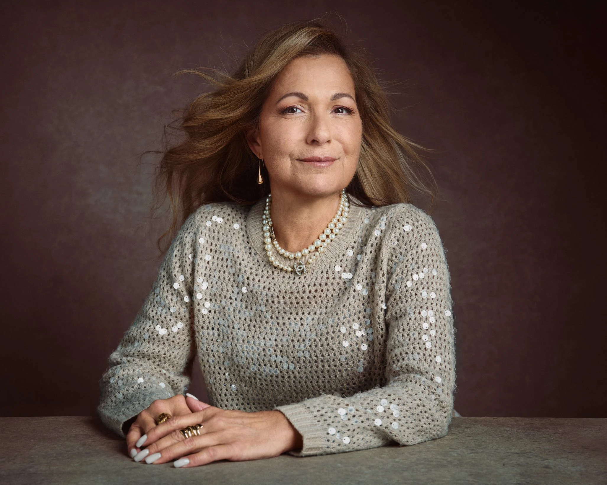 A middle-aged woman with light brown hair styled in loose waves, wearing a pearl necklace, gold earrings, a beige knit sweater with sequins, and gold rings, sitting at a table with her hands folded in front of her, looking confidently at the camera a