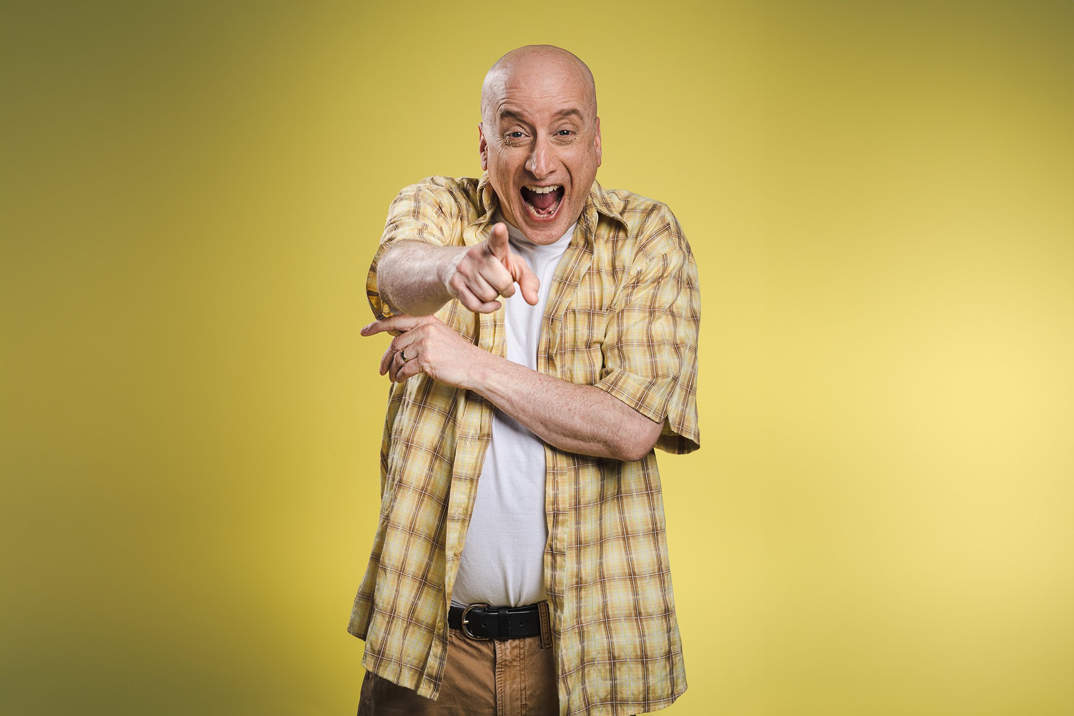 A bald man wearing a yellow plaid shirt and beige pants is pointing at the camera and smiling broadly against a yellow background.