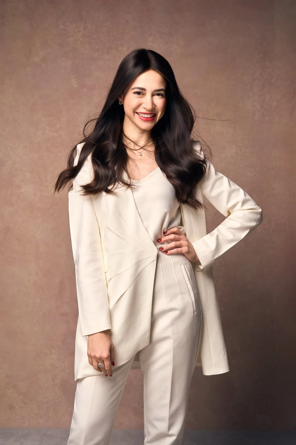 A woman with long dark hair in a cream suit, smiling and standing with one hand on her hip, against a brown textured background.