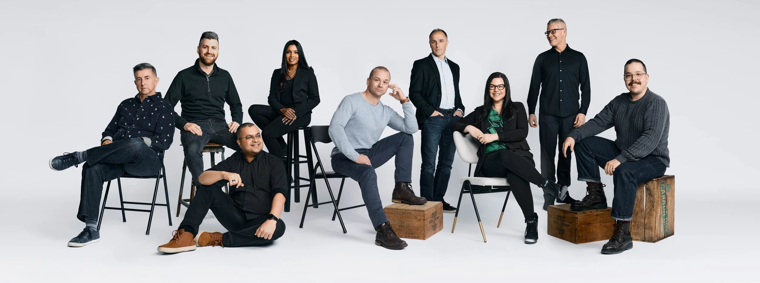 A diverse group of ten people posing together in a studio with a white background, sitting and standing on various chairs and wooden boxes.