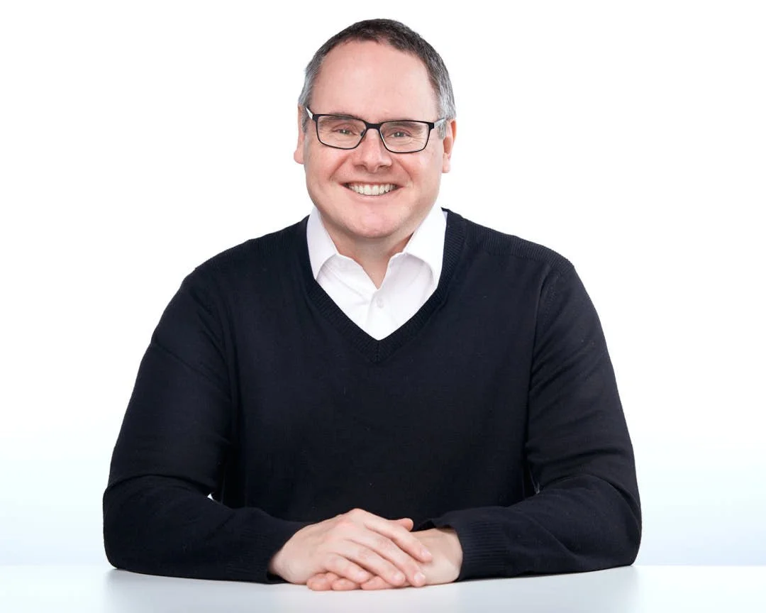 Smiling man with glasses and a white shirt under a black sweater, posed with hands on the table — clean, approachable corporate headshot for Oakville executives and team leaders.
