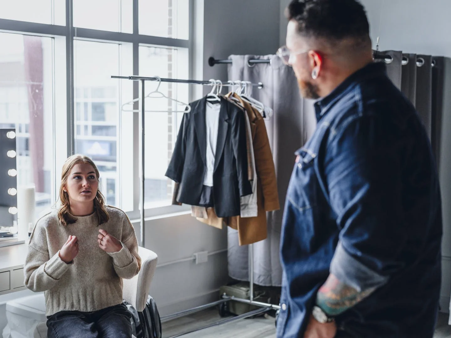 A woman with shoulder-length hair wearing a beige sweater talking to a headshot photographer with tattoos wearing a denim jacket in an office or studio with large windows and a clothing rack with jackets and shirts.