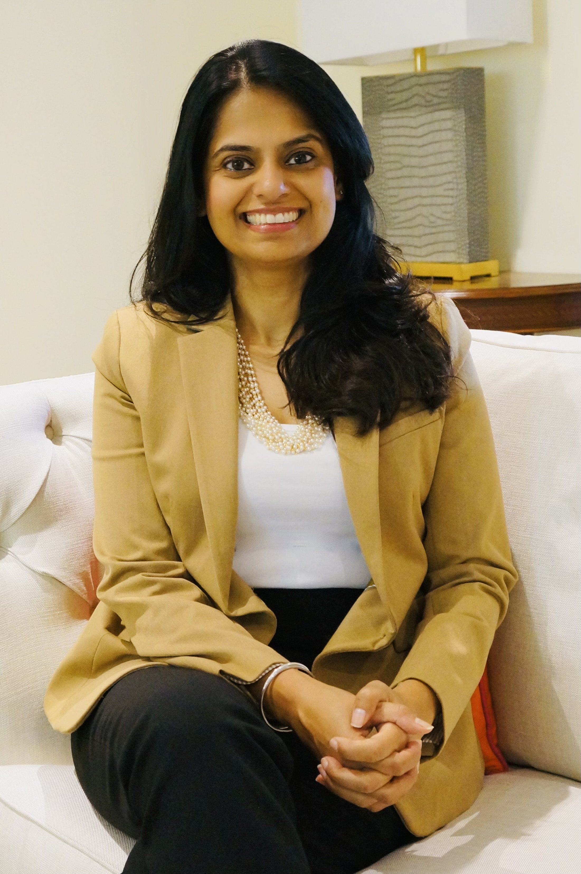 A woman sitting on a white couch, wearing a beige blazer, white blouse, and black pants. She has long dark hair and is smiling, with hands clasped in her lap. A lamp on a table is visible in the background.
