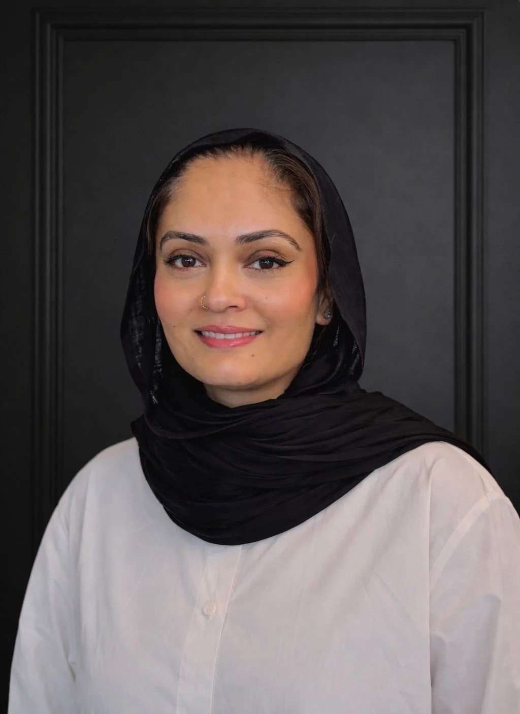 A woman with light skin, wearing a black headscarf and a beige shirt, stands in front of a dark, textured background. She has a friendly expression with a slight smile.