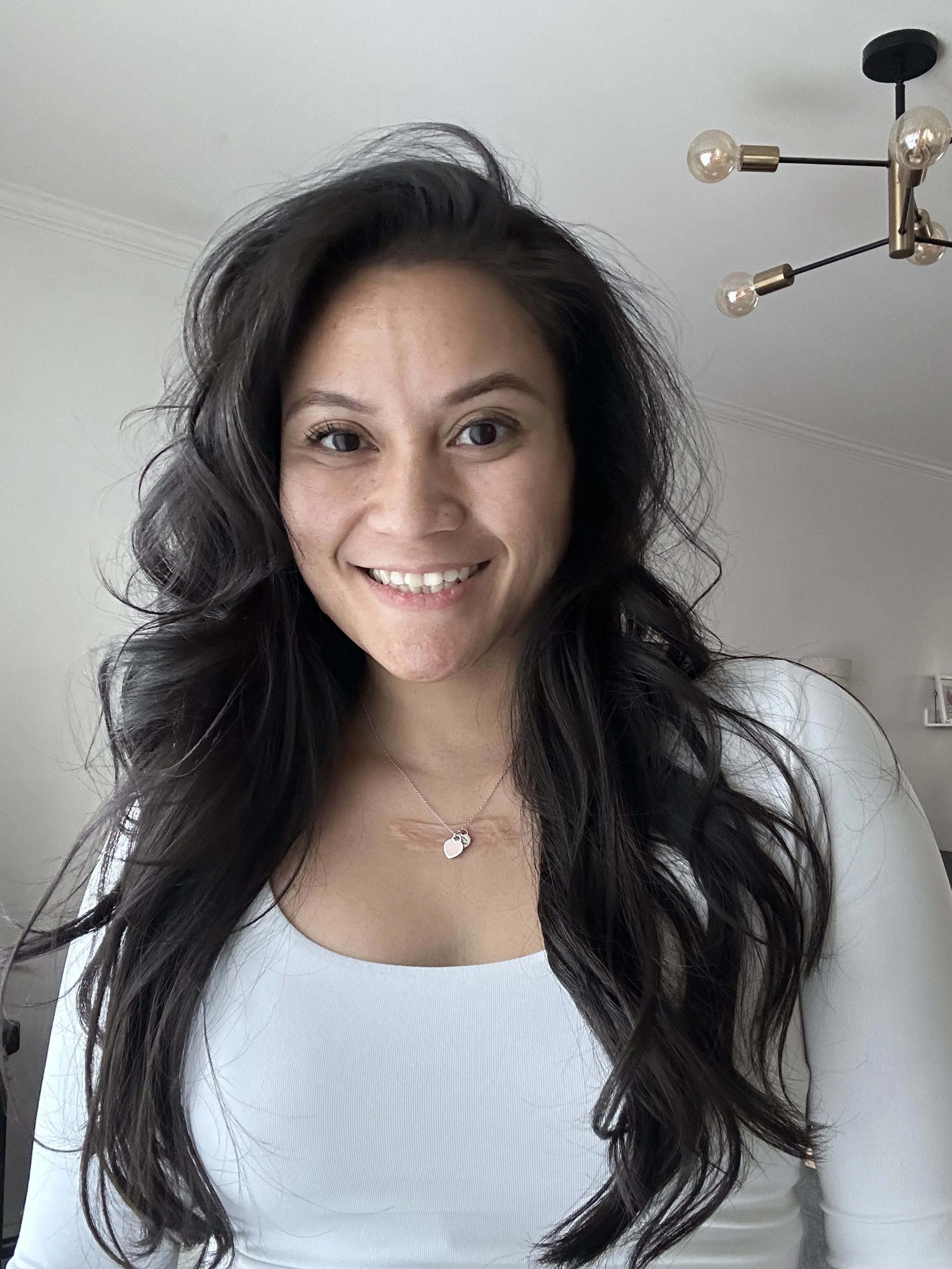 A woman with long, wavy black hair smiling in a well-lit room, wearing a white top and a necklace with a small heart-shaped pendant.