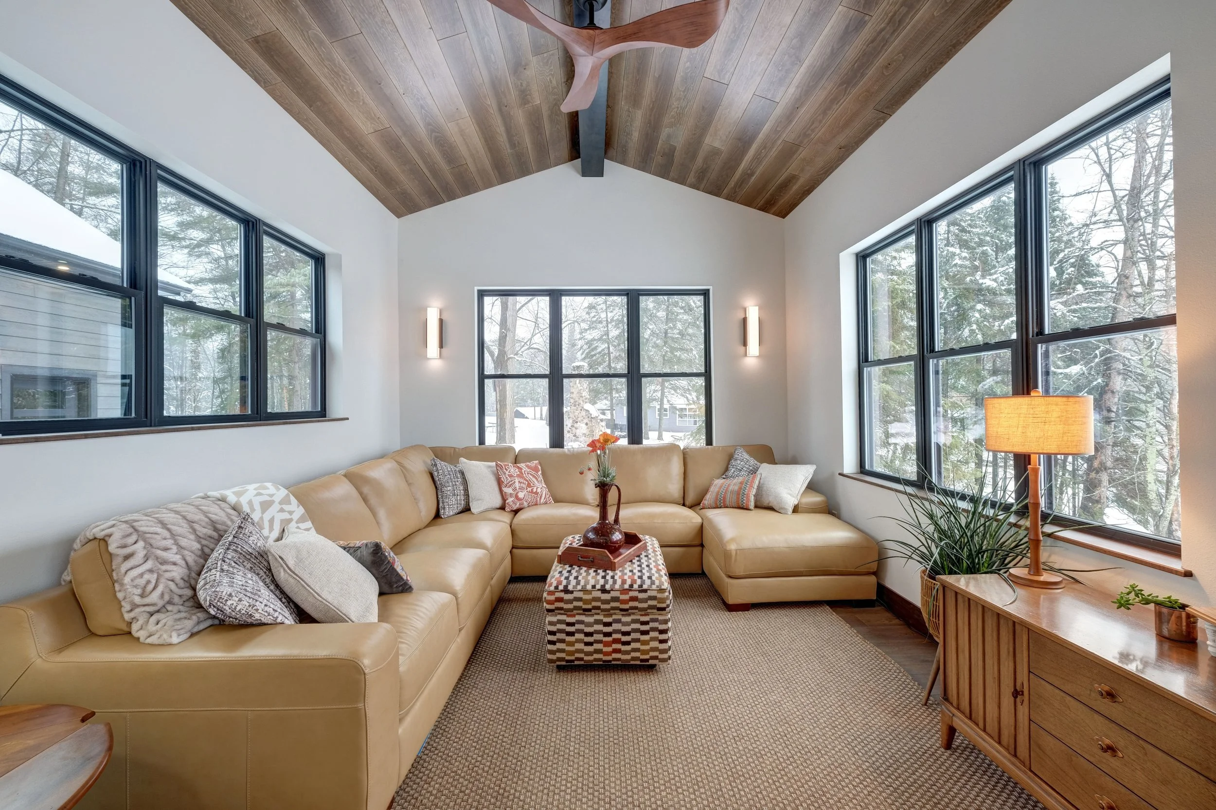 Living room with large beige sectional sofa, wooden ceiling, and three large windows showing snowy trees outside.