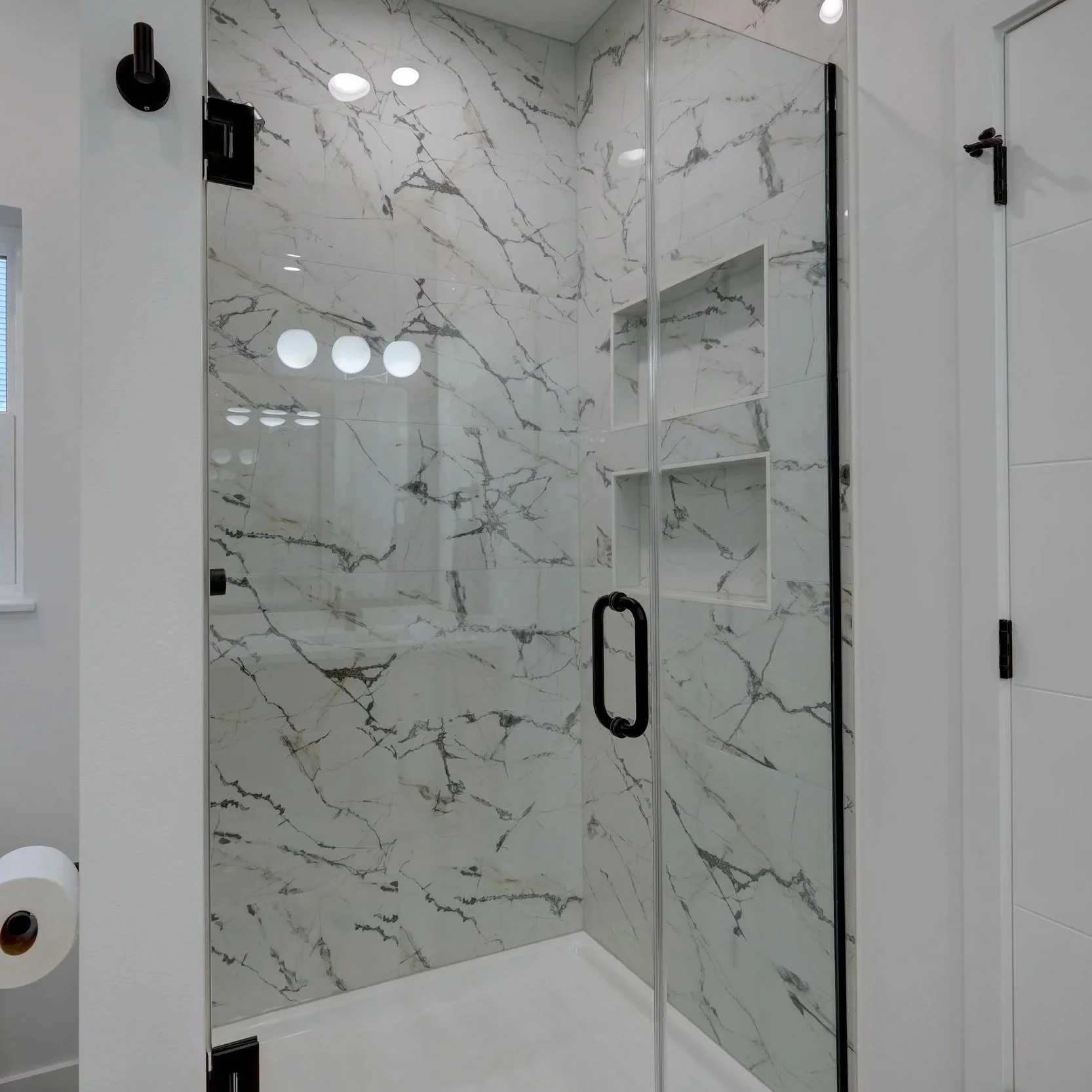 A modern bathroom with a glass shower enclosure featuring white marble walls with gray veining and built-in shelves.