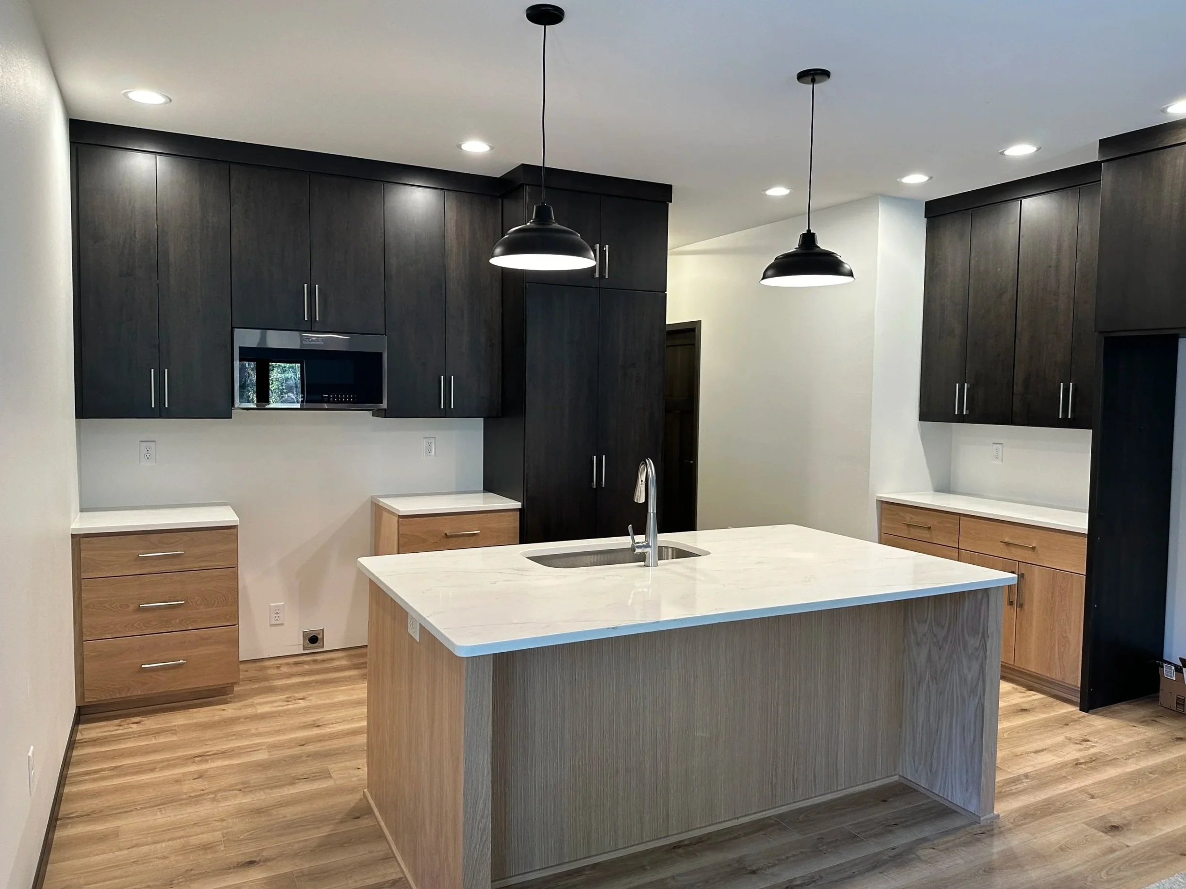 Modern kitchen with dark upper cabinets, light lower cabinets, a central island with a sink, light wood flooring, white countertops, and hanging black pendant lights.