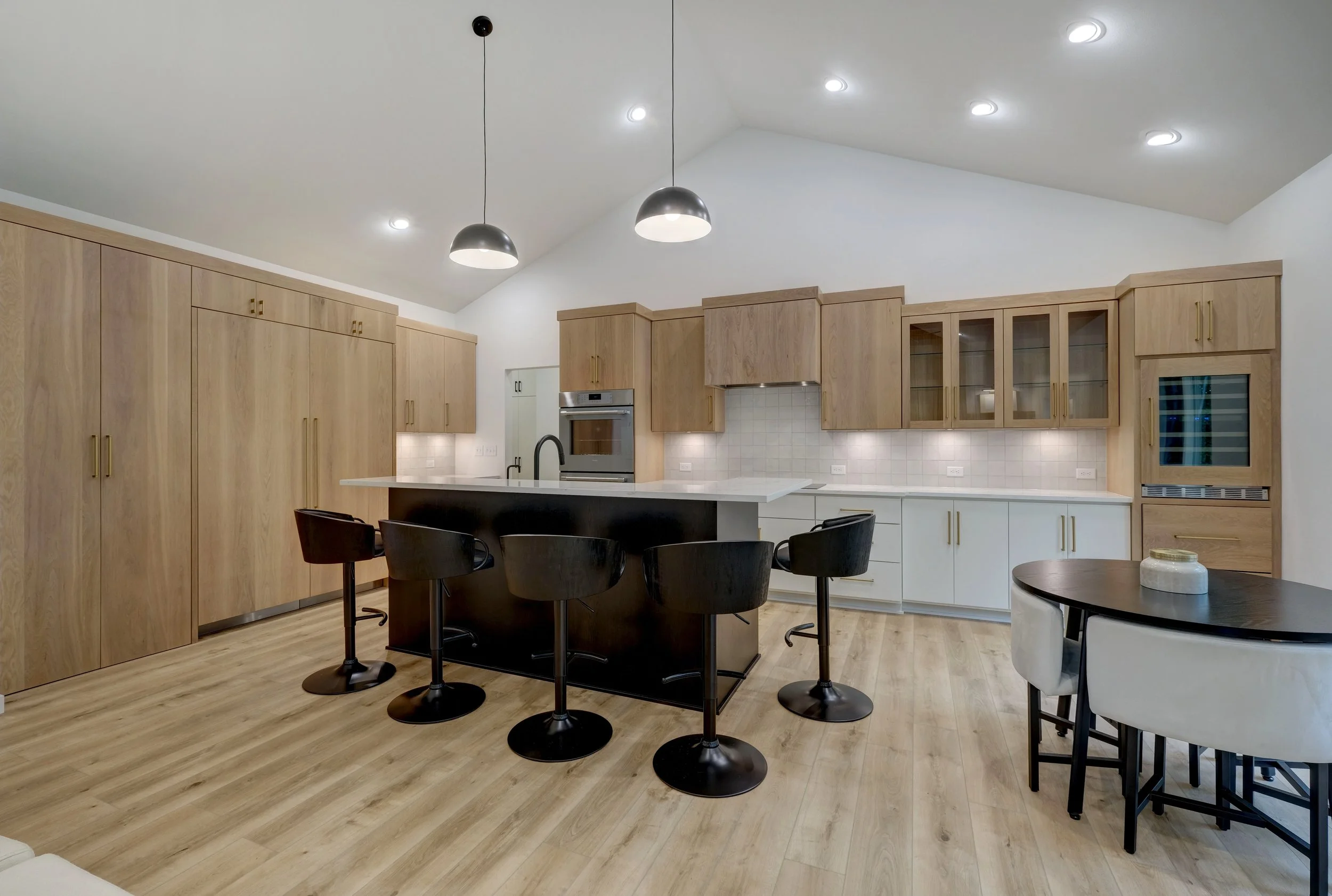 Modern kitchen with light wood cabinets, an island with black base and black bar stools, white countertops, and a round dining table with white chairs.