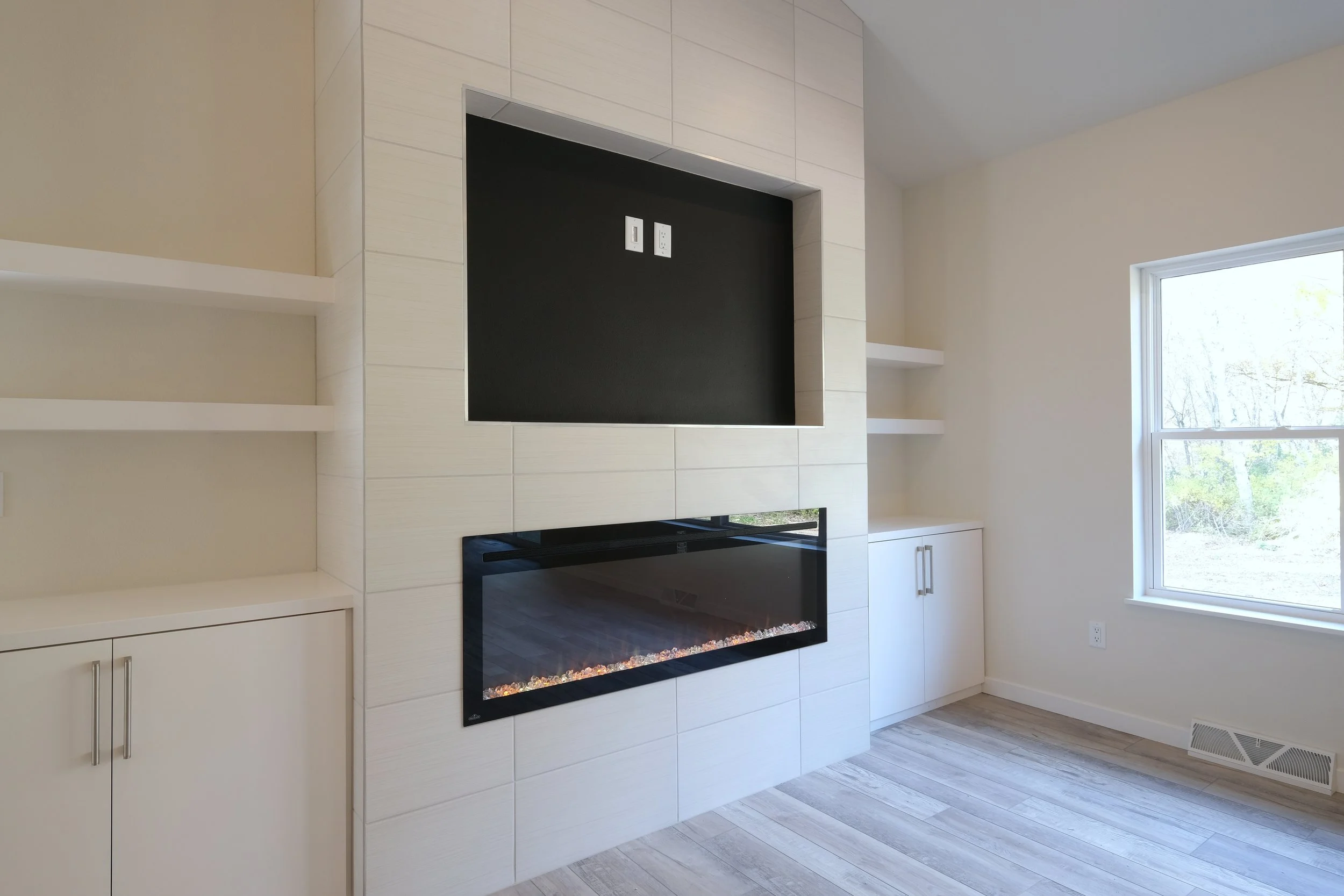 Empty living room with a modern white built-in fireplace and black accent behind it, white shelves on either side, and a large window to the right showing outdoor trees.
