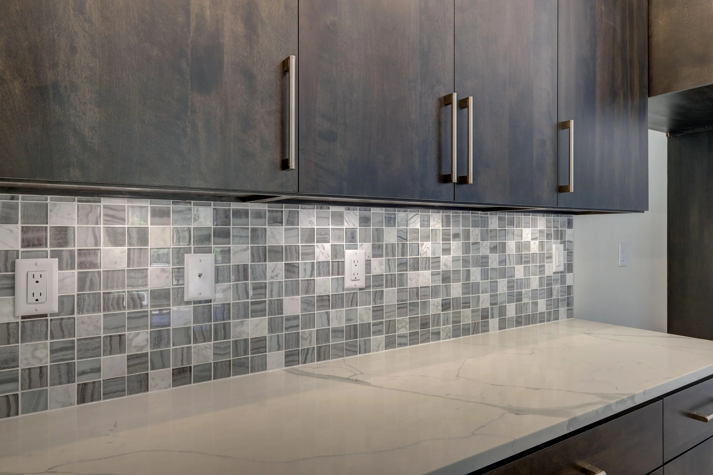 Close-up of kitchen backsplash with small gray and white tiles, dark wood cabinets with long silver handles, and a light-colored marble countertop with subtle veining.