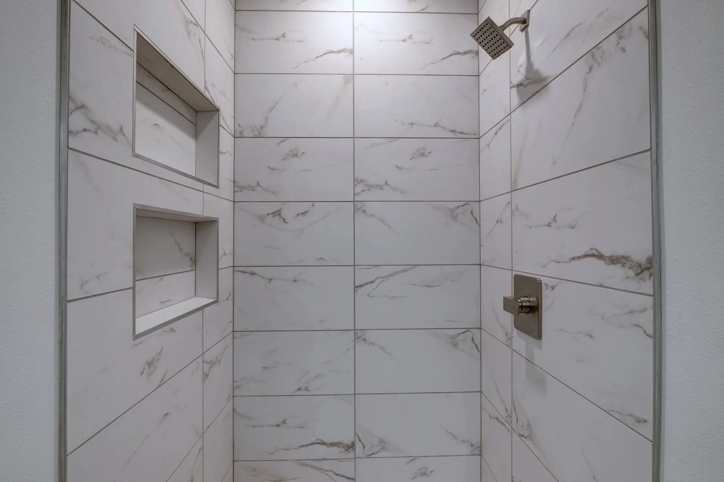A shower with large white marble-patterned tiles, steel fixtures, a square overhead showerhead, two built-in tiled shelves, and a gray door frame.