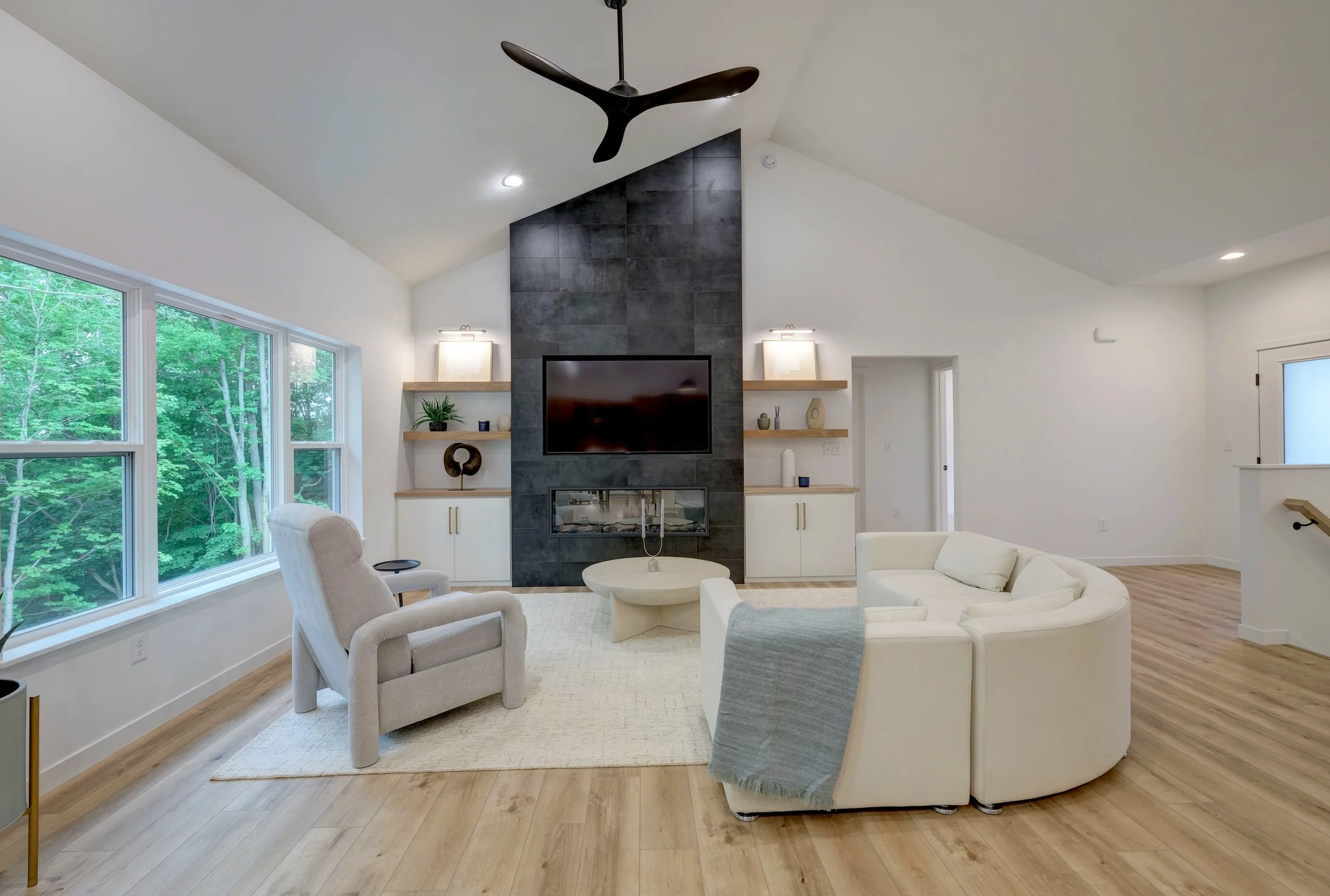 Living room with large windows, white walls, a black ceiling fan, a modern fireplace with a flat-screen TV above, built-in shelves with decorations, white curved sofa with a blanket, beige armchair, wooden flooring, and minimalistic decor.
