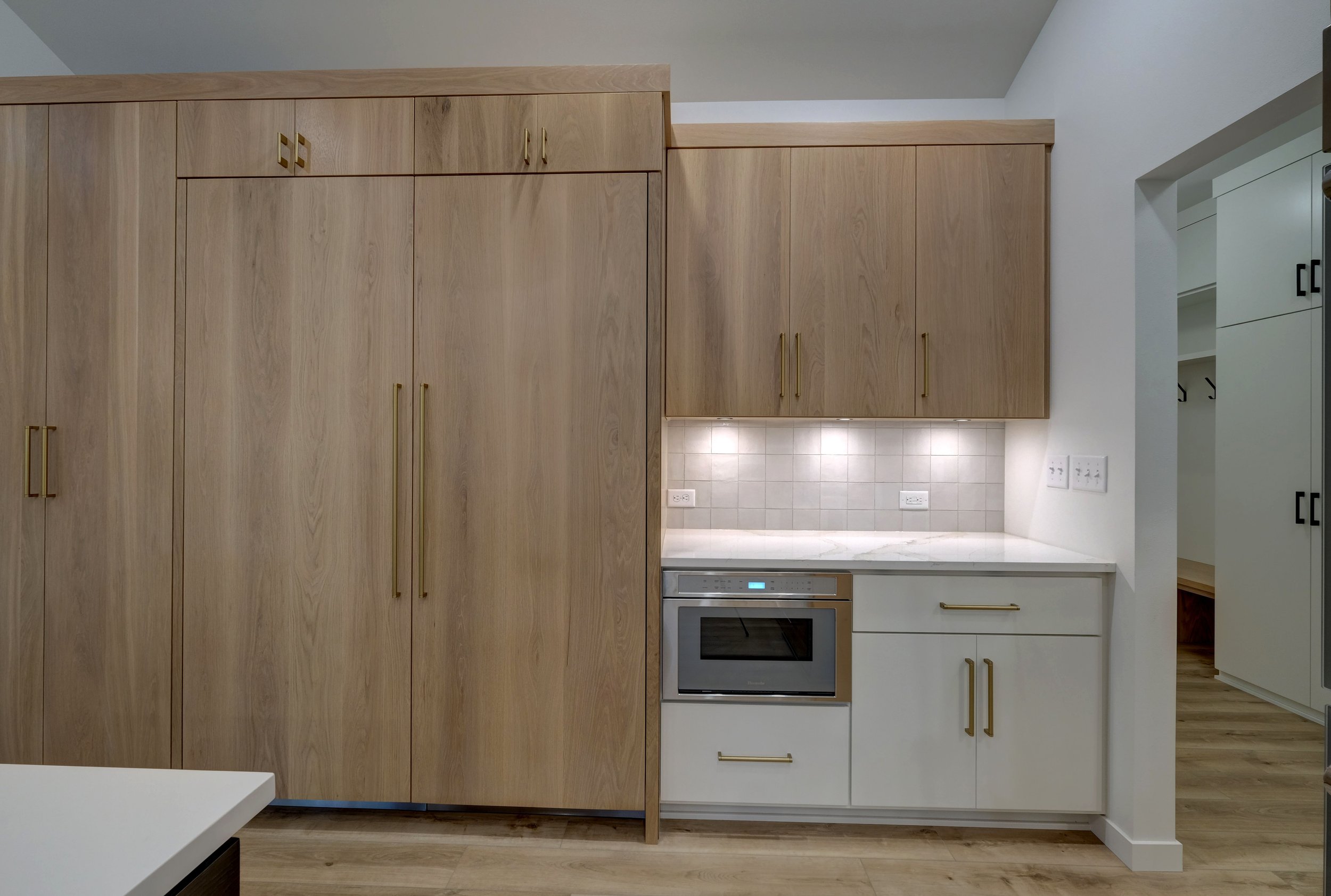 Modern kitchen with light wood cabinets, white countertop, tiled backsplash, built-in microwave, and under-cabinet lighting.