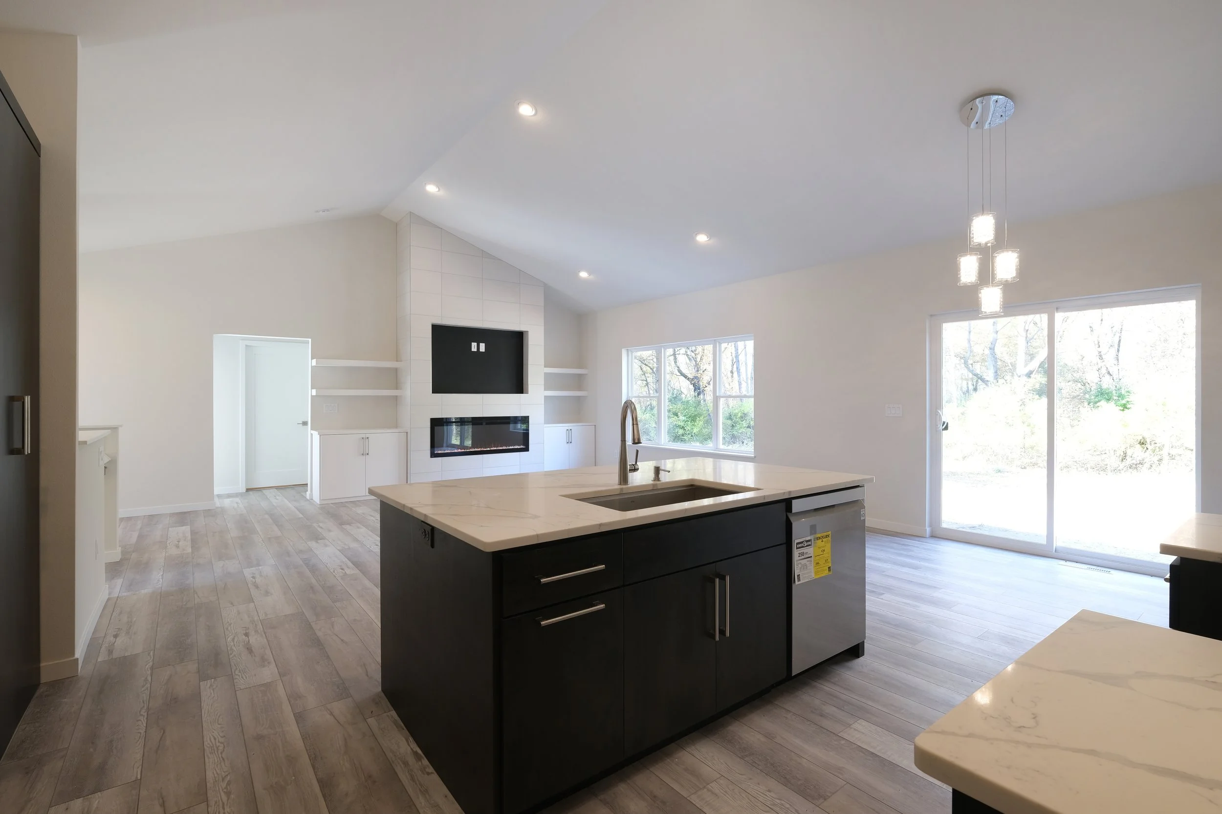 Open-concept kitchen and living room with white walls, light wood floors, kitchen island with black cabinetry, modern pendant lights, large windows, sliding glass door, fireplace, and built-in shelves.