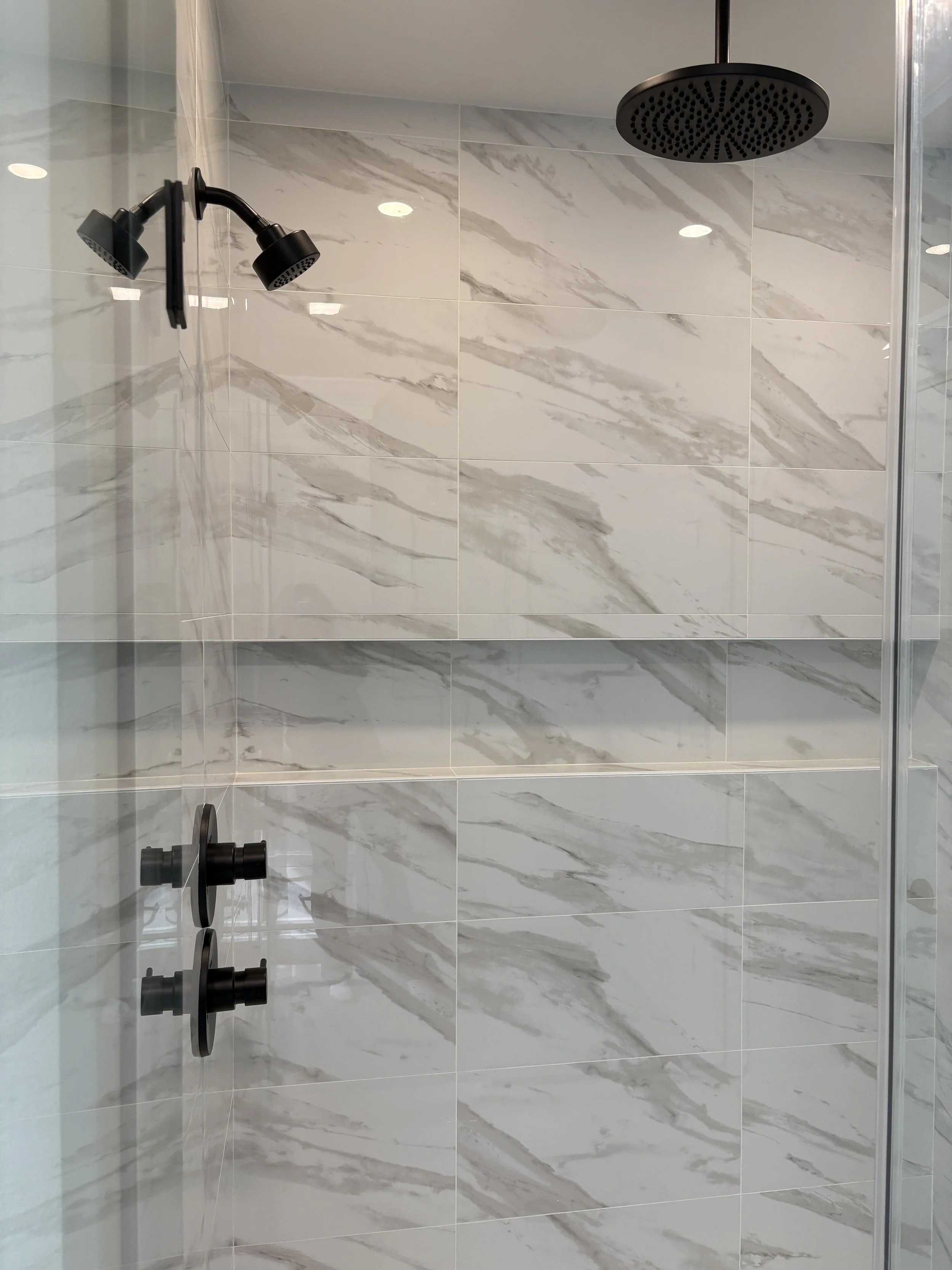 Modern shower with black fixtures, a rain shower head, and white marble-like tiles with gray veins on the walls.
