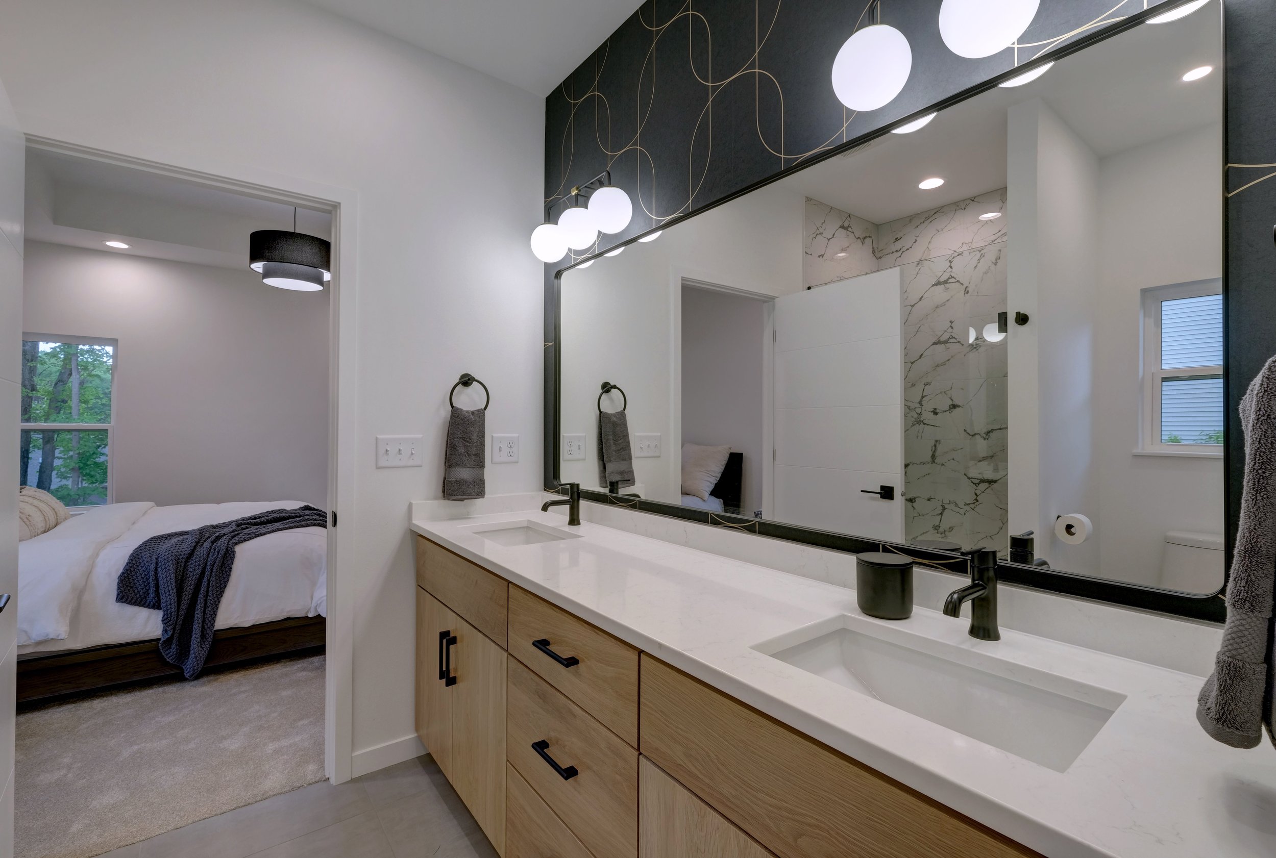 Modern bathroom with double sink vanity, large mirror, black fixtures, white countertop, and a view into a bedroom with a bed, window, and ceiling light