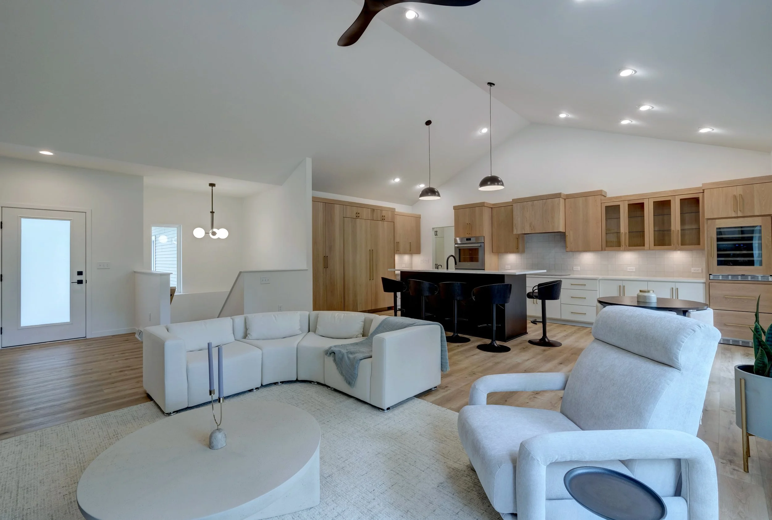 Open-concept living room and kitchen with white sofa, beige armchair, light wood cabinets, black kitchen island with bar stools, and modern lighting fixtures.