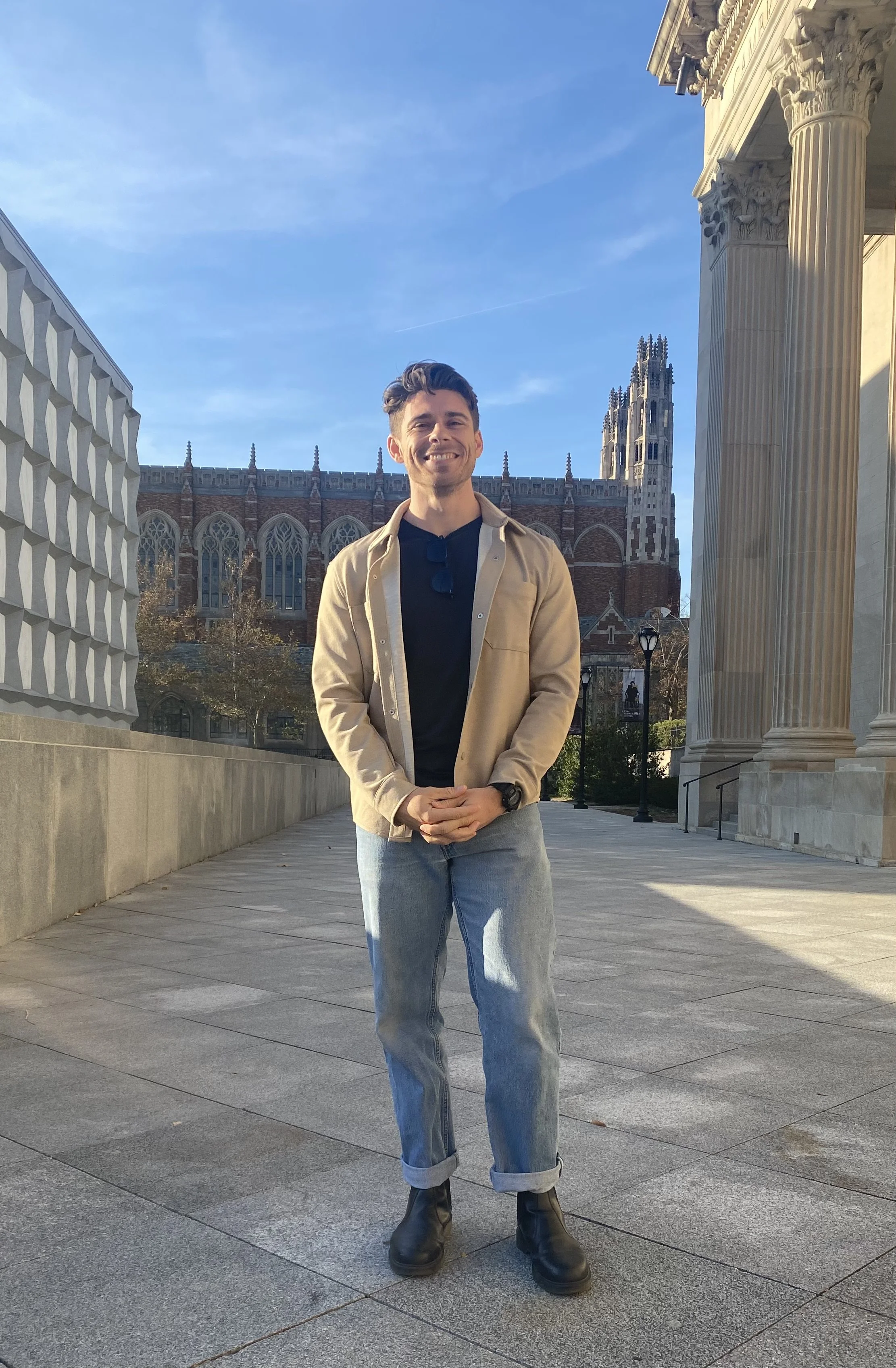Person standing outside near architectural columns and buildings, smiling, dressed in casual attire with a jacket and jeans, under a clear blue sky.