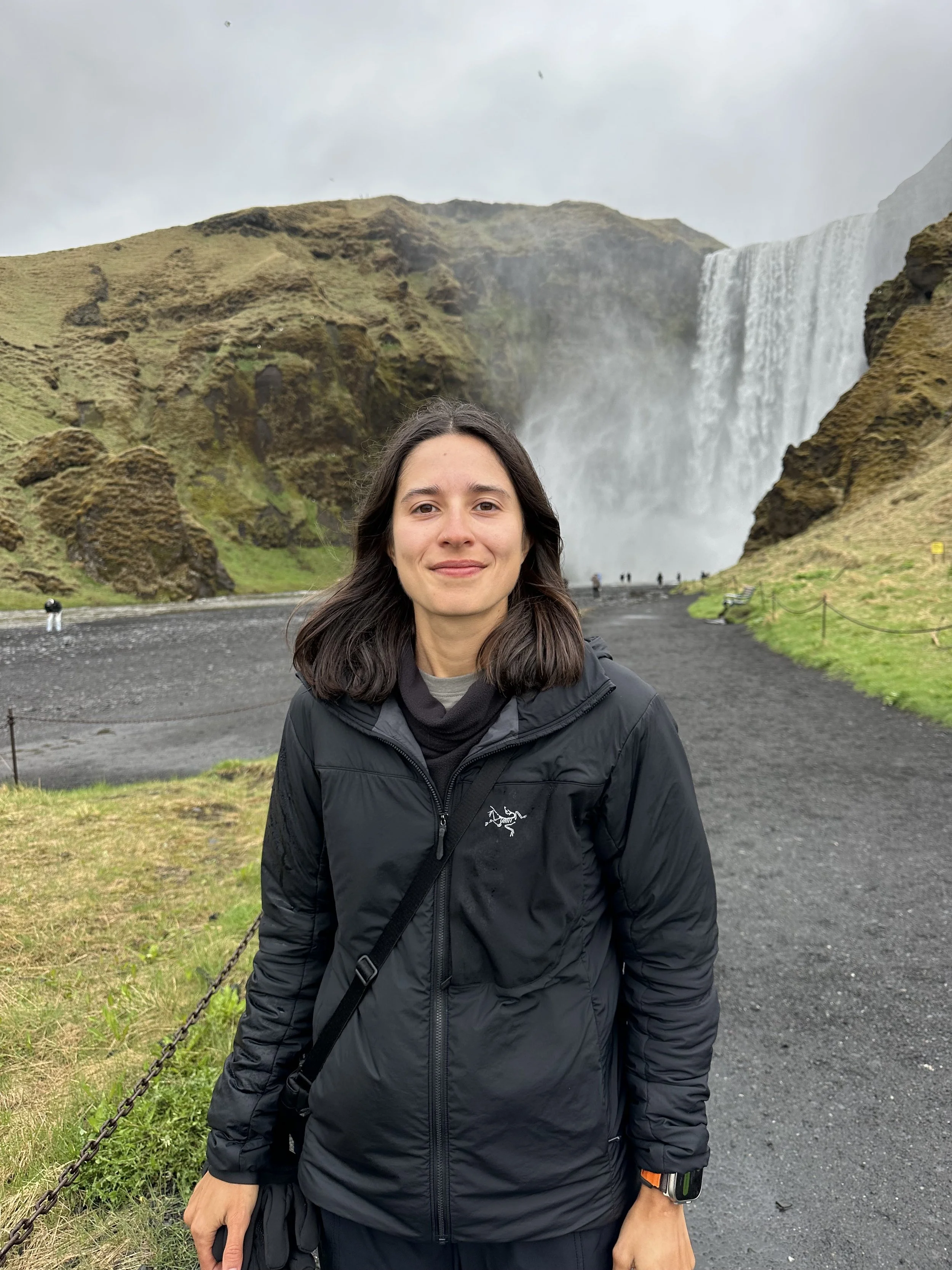 Person standing in front of a large waterfall, wearing a black jacket, with grassy hills and a cloudy sky in the background.