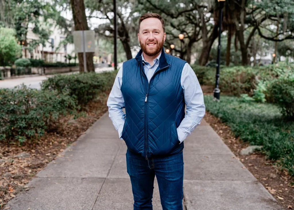 Man wearing a blue vest and jeans standing on a sidewalk in a park with trees in the background.
