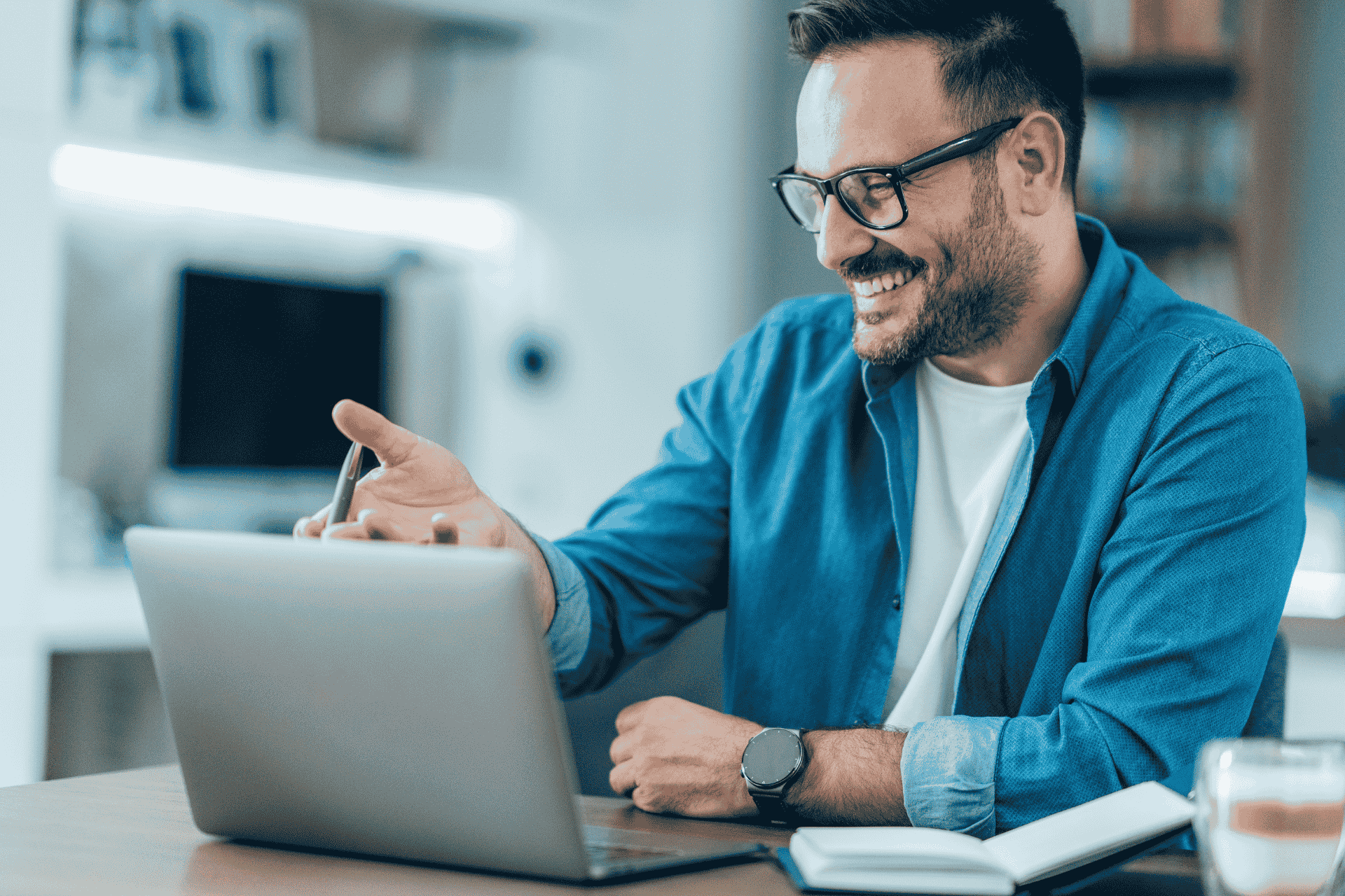 A smiling man holding a pen sitting in front of laptop with his hand up showing his is happy and agreeing with the conversation he is in with a video call.