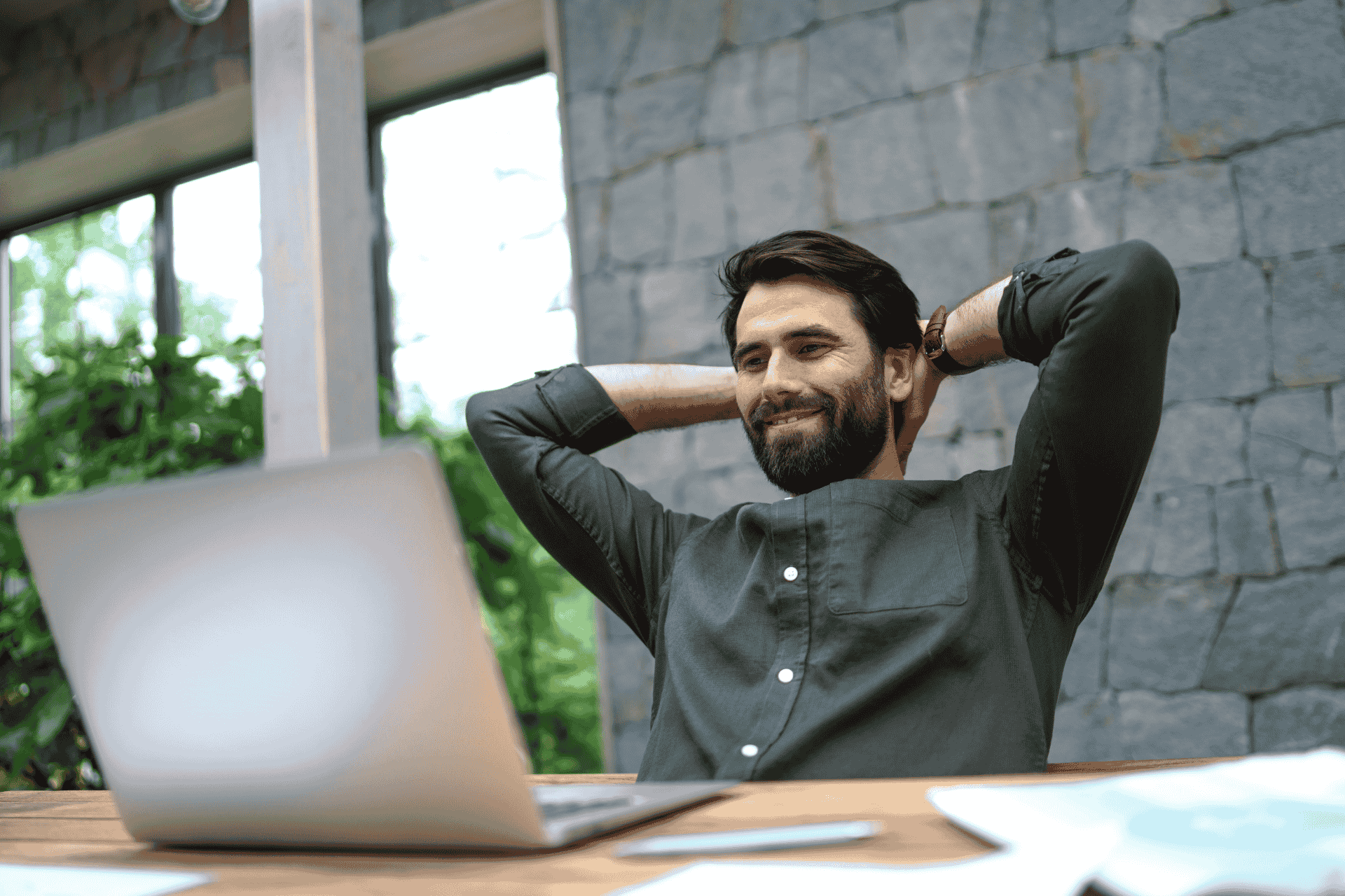 A bearded man sitting at table in front of laptop with his hands and arms behind his head and a big smile showing he is confident and happy with results he is getting in business.