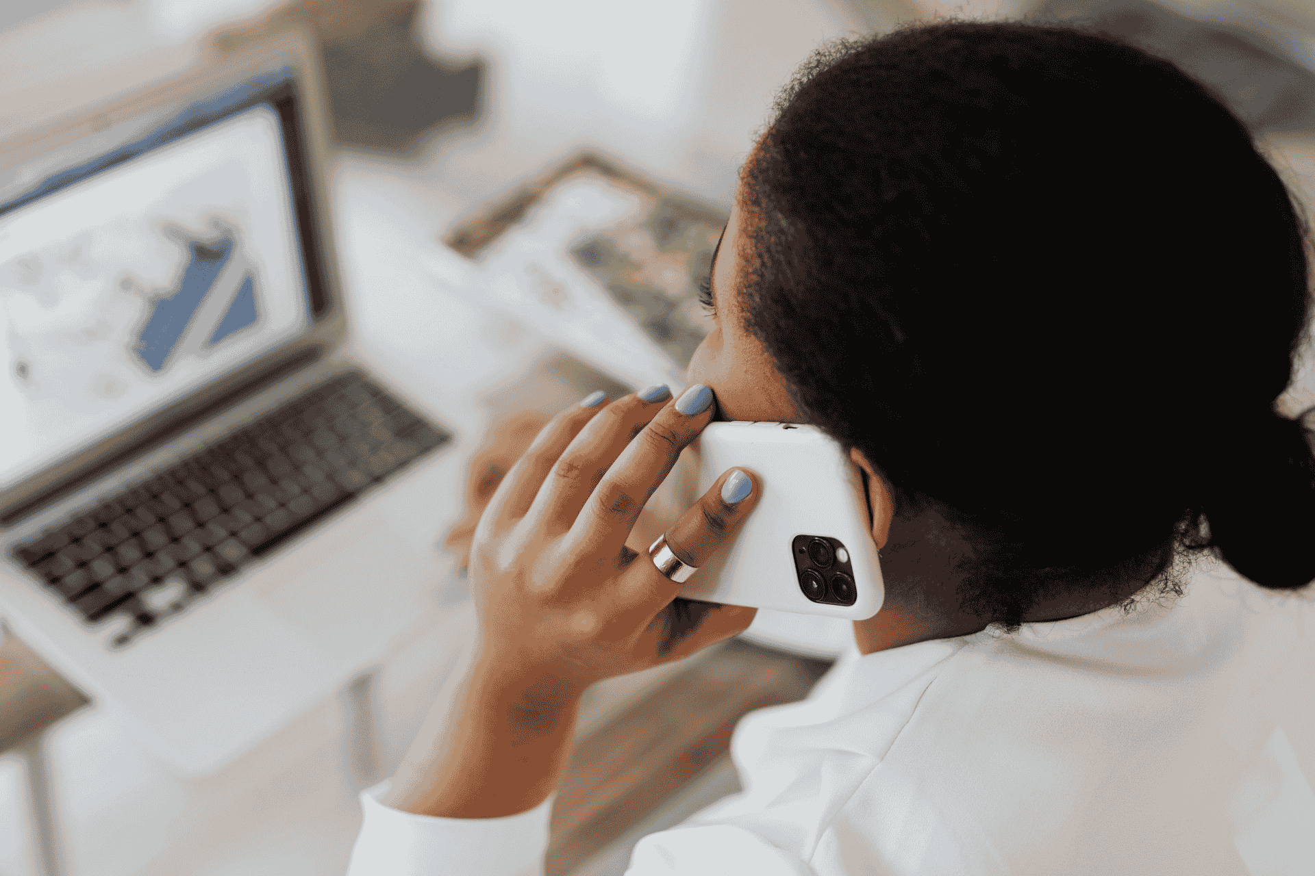 A woman faces a laptop while holding a cell phone to ear showing she is having a business discussion.