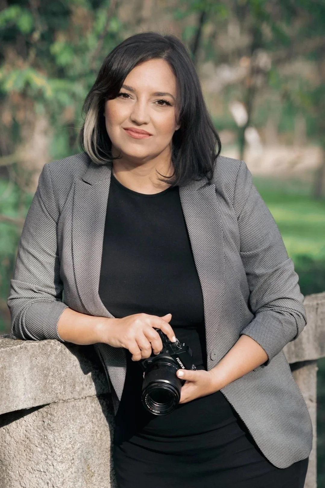A woman with dark hair and a grey blazer holding a camera outdoors, standing near a stone ledge, with green trees in the background.