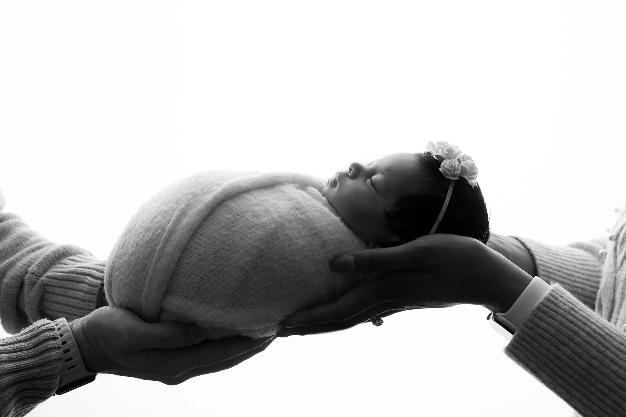 A tender newborn family moment: Mother and father's hands cradling their swaddled baby boy in a soft, airy studio setting.